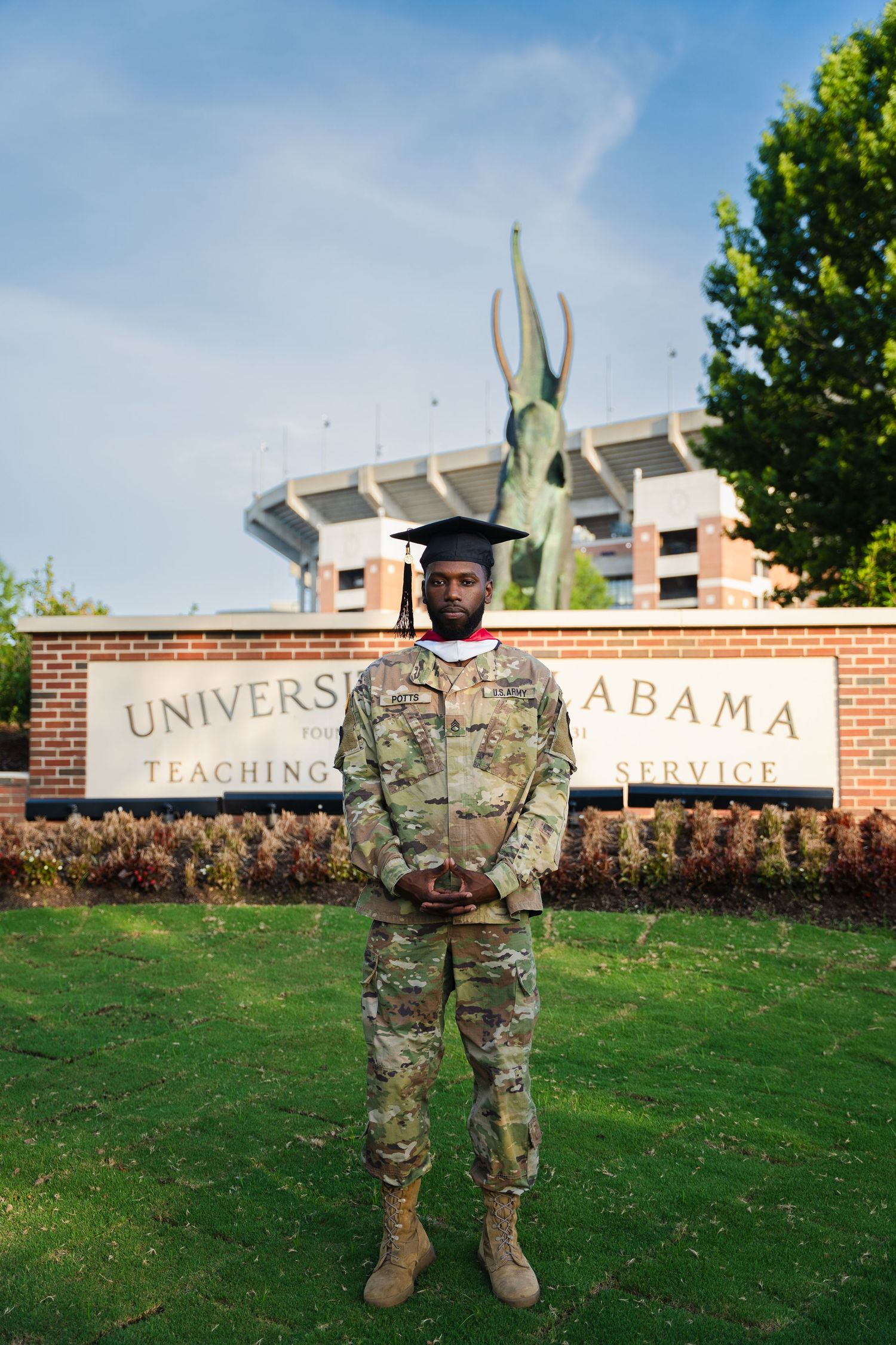 Military service member in camouflage uniform stands in front of University of Alabama entrance sign.