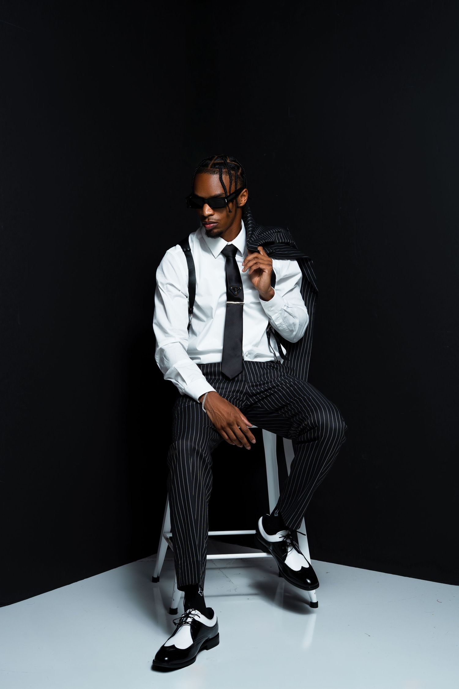 Stylish figure in white dress shirt, striped pants and suspenders poses confidently on stool against dark background.