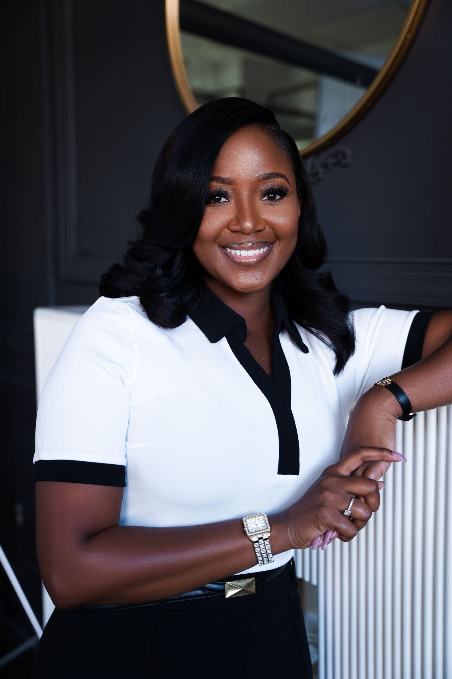 Professional woman in white polo shirt with black trim smiles confidently against dark wall with round mirror.