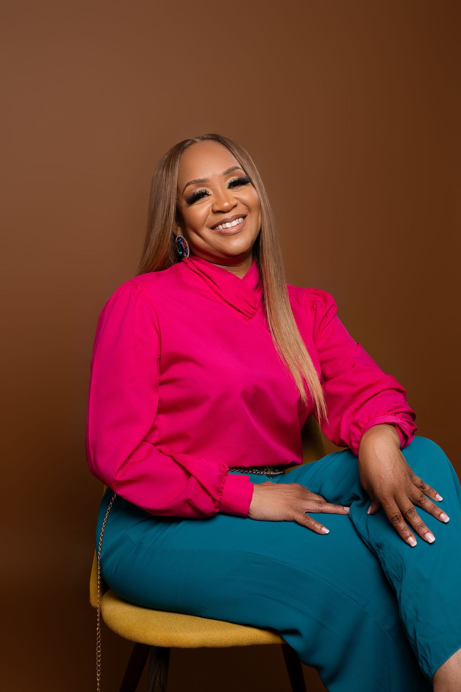 Person in bright pink top and teal pants poses on chair against brown background.