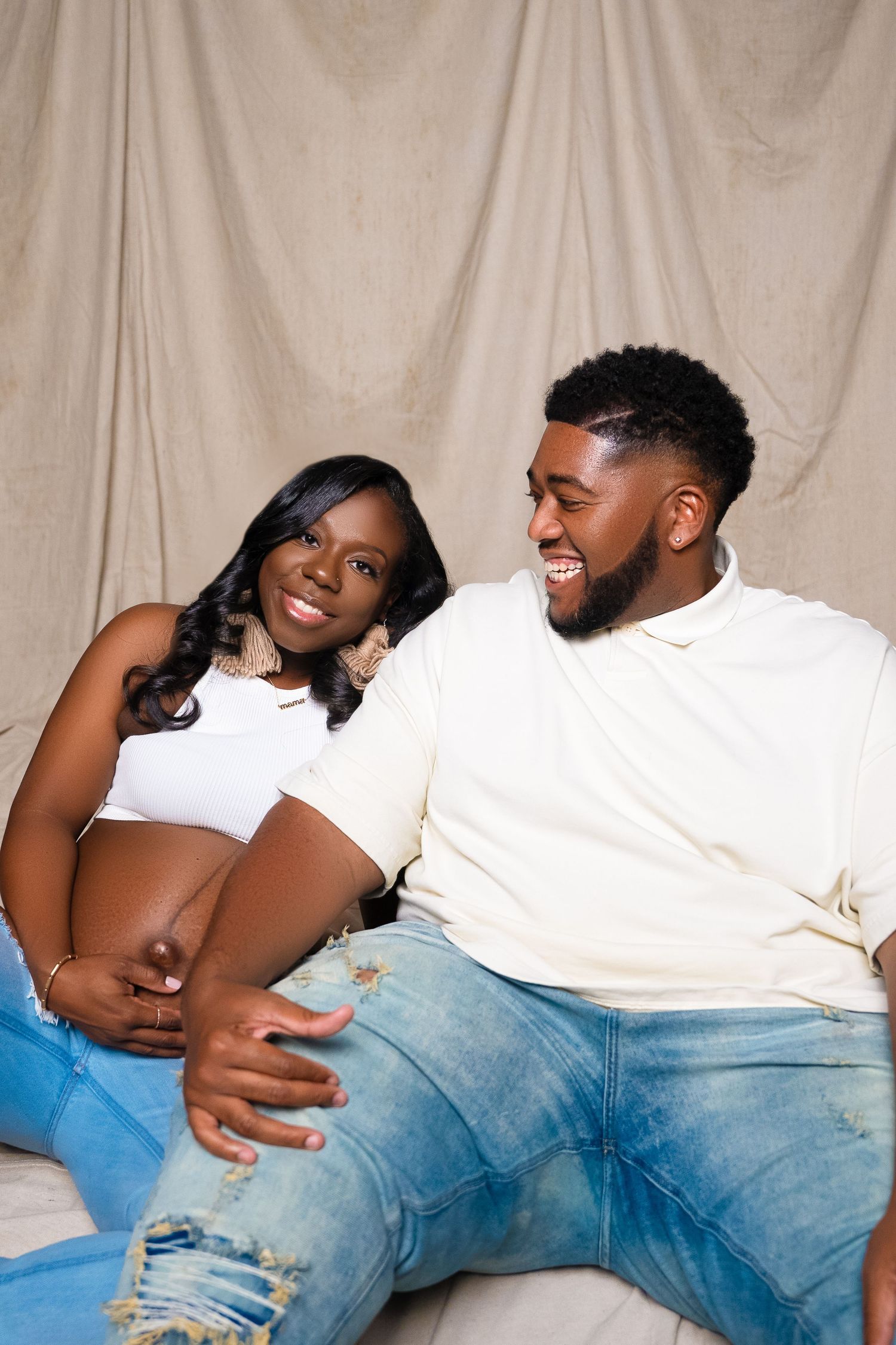Casual maternity photo shoot with couple in white and denim against beige backdrop.