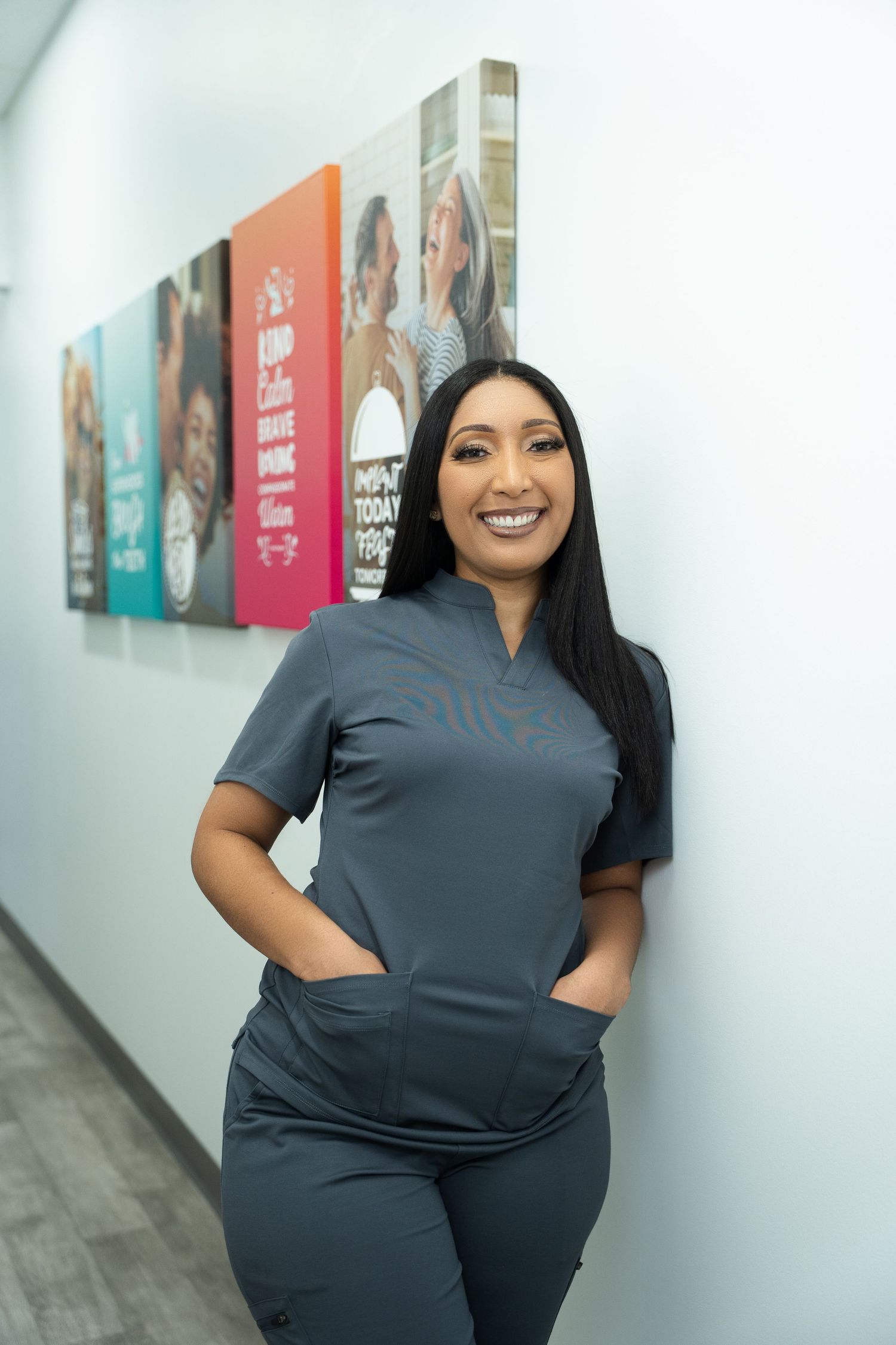 Healthcare worker in gray scrubs smiles while posing against office wall with colorful artwork display.