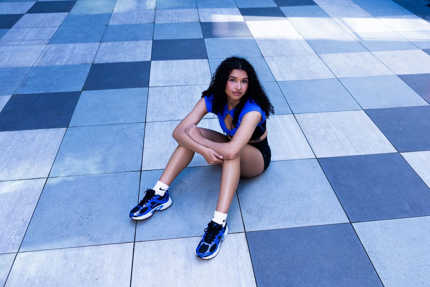 Young woman in blue sneakers and shorts sitting on a checkered tile floor outdoors.