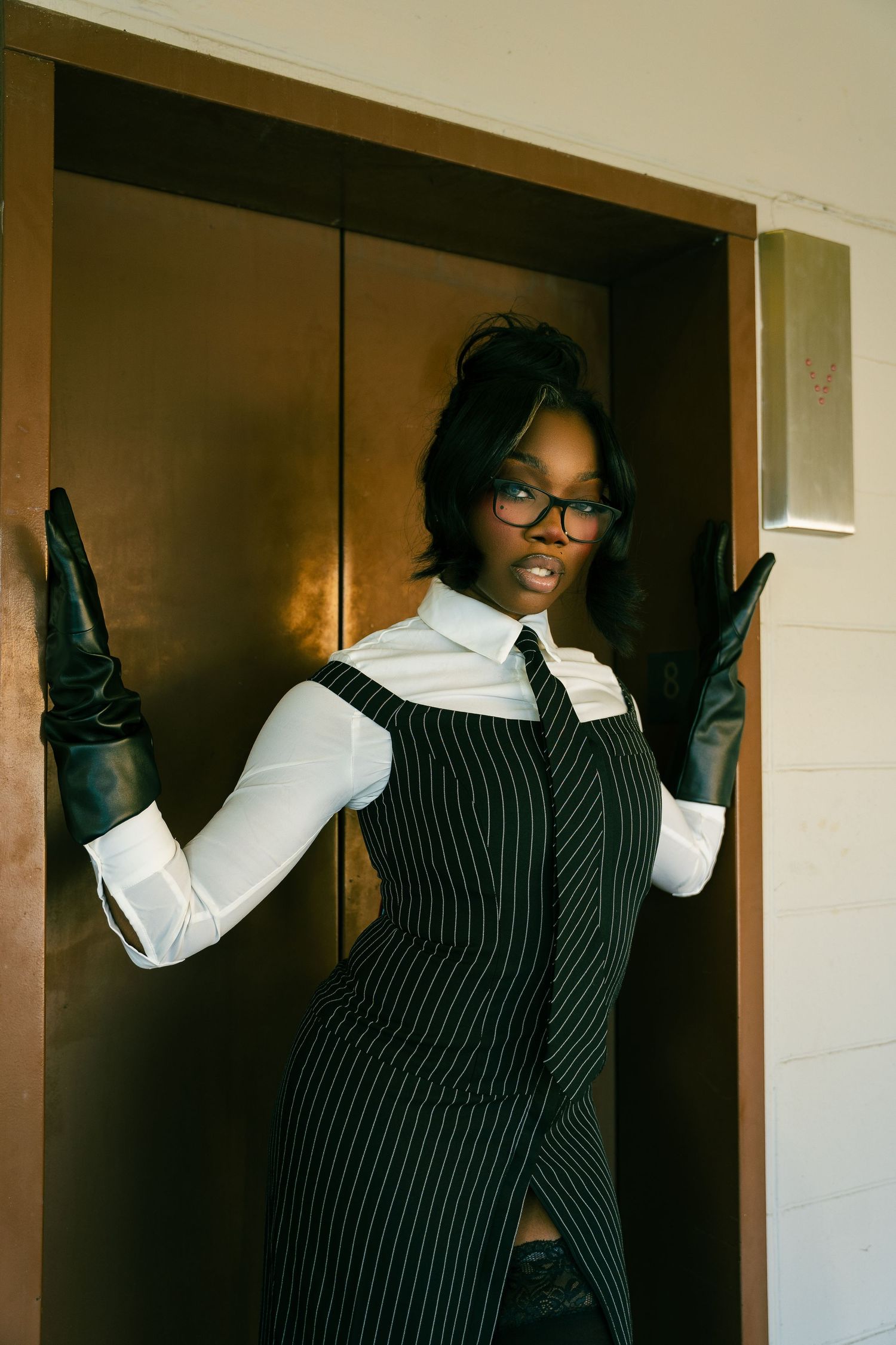 Person wearing a pinstriped dress, white collared shirt, glasses and long black gloves poses in a doorway.