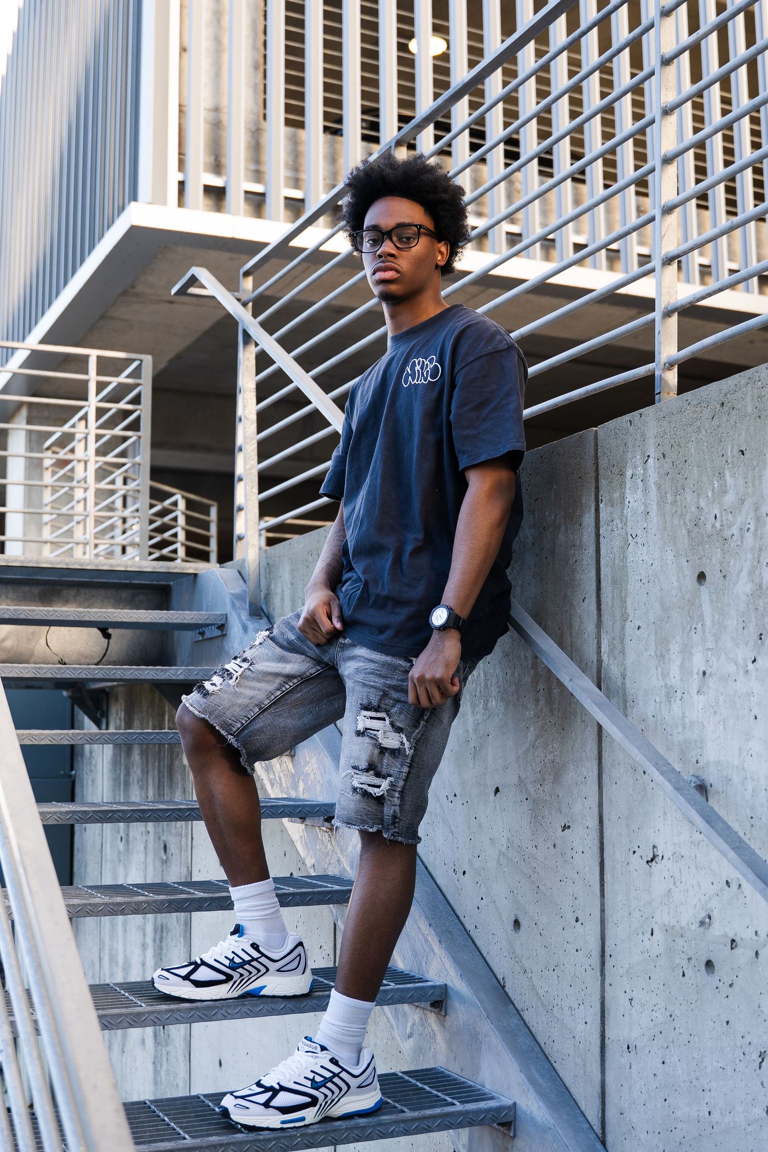 Urban fashion shot of person in navy t-shirt and cargo shorts posing on concrete stairs.