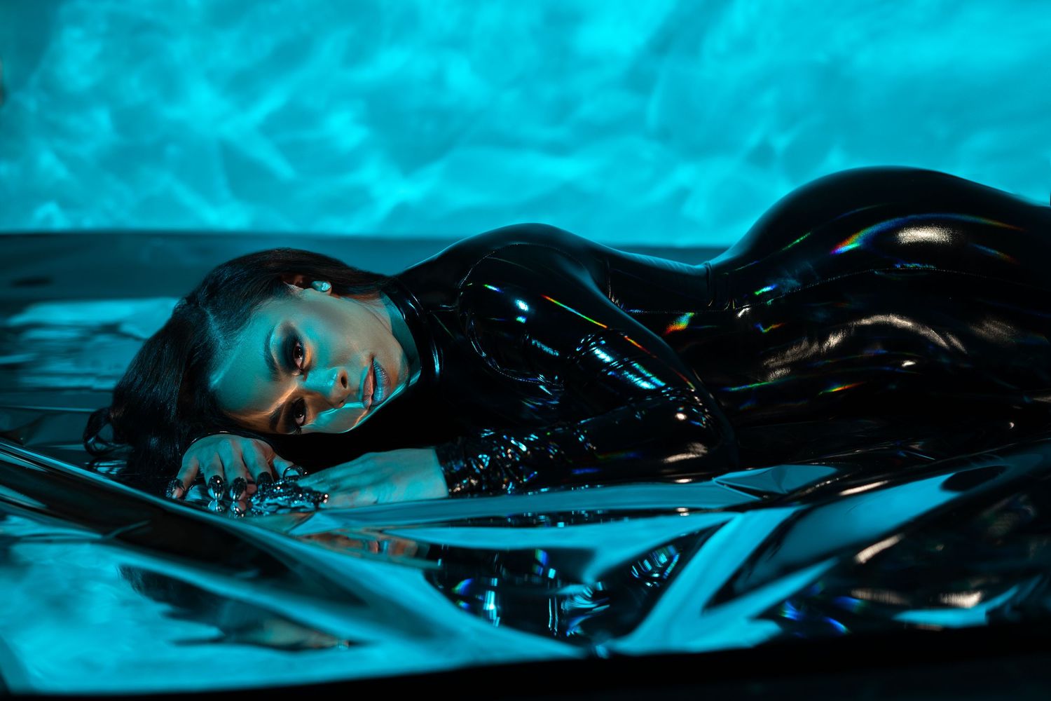 Artistic fashion photo in shiny black latex with dramatic turquoise lighting and reflective surfaces.