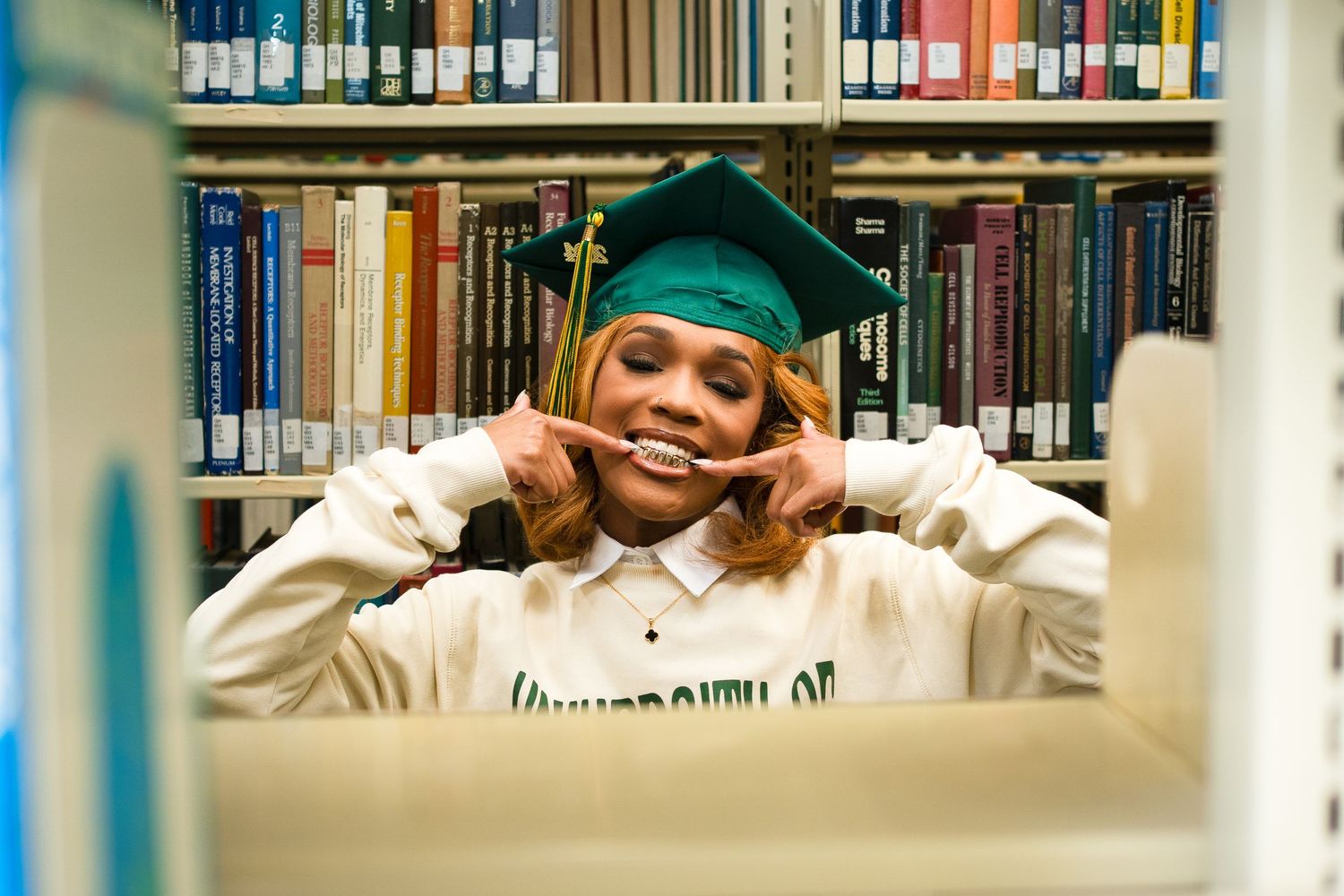 Graduate in green cap peeking through library bookshelves with playful expression.
