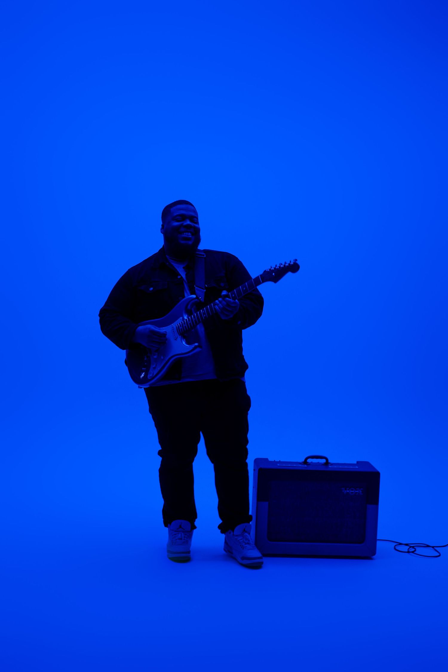 Guitar player stands with instrument and amplifier in dramatic blue lighting.