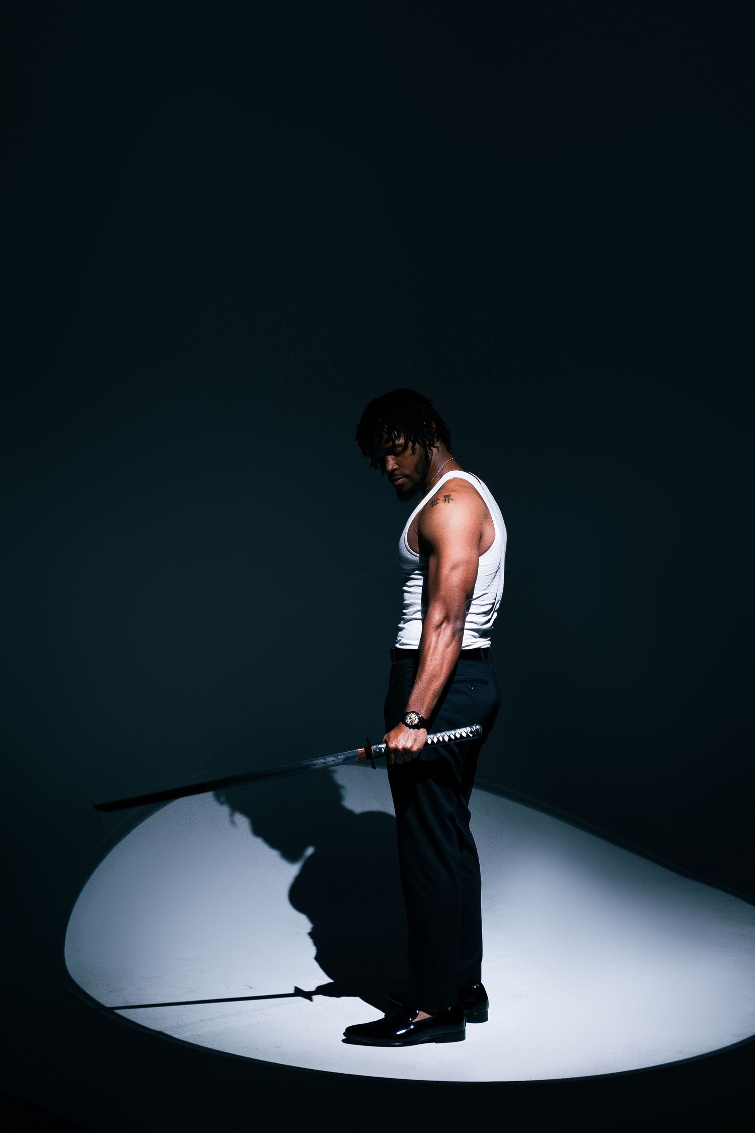 Athletic figure in white tank top stands in dramatic spotlight holding weights.