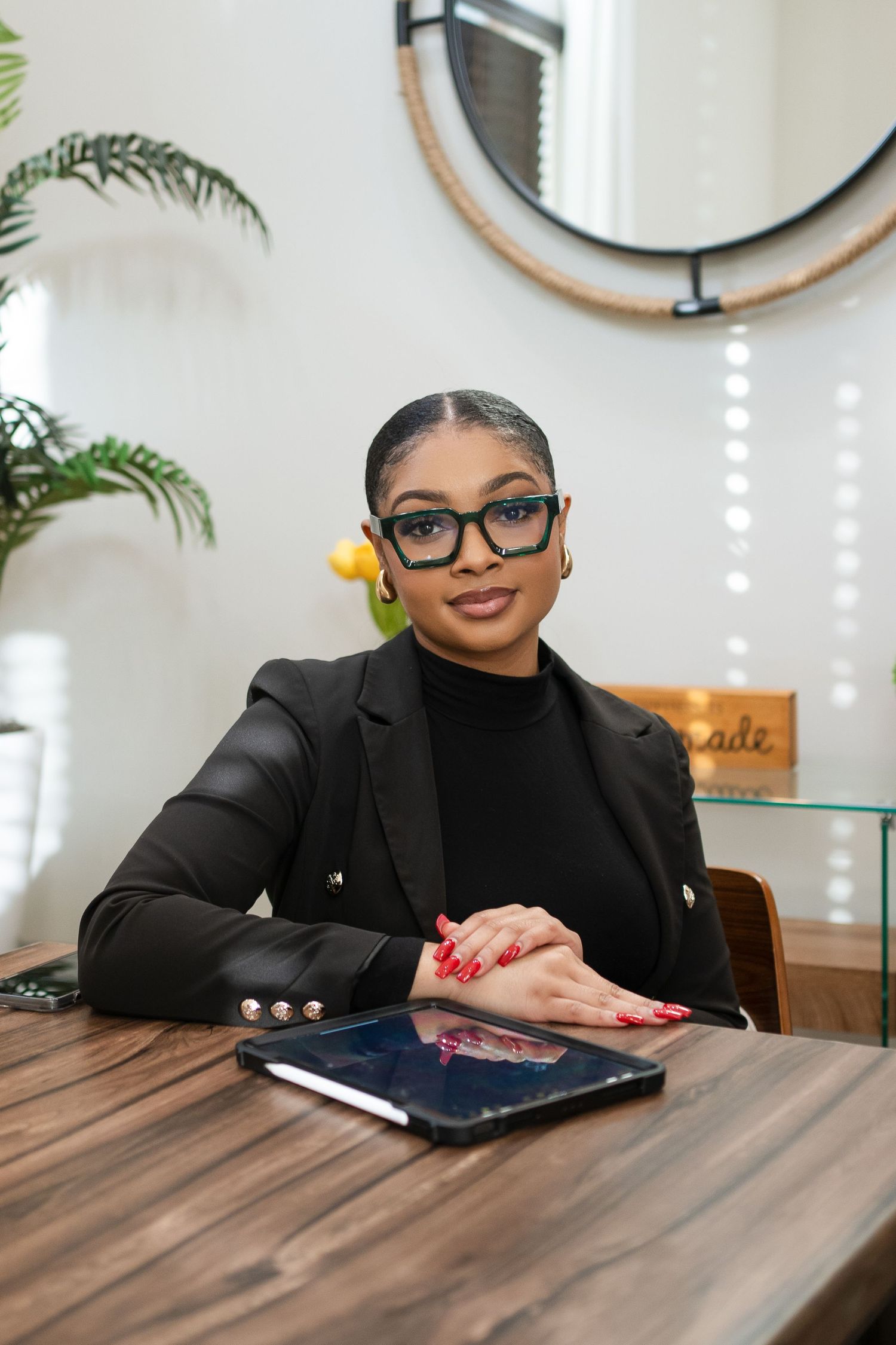Professional seated at wooden desk wearing black blazer and glasses in modern office setting.