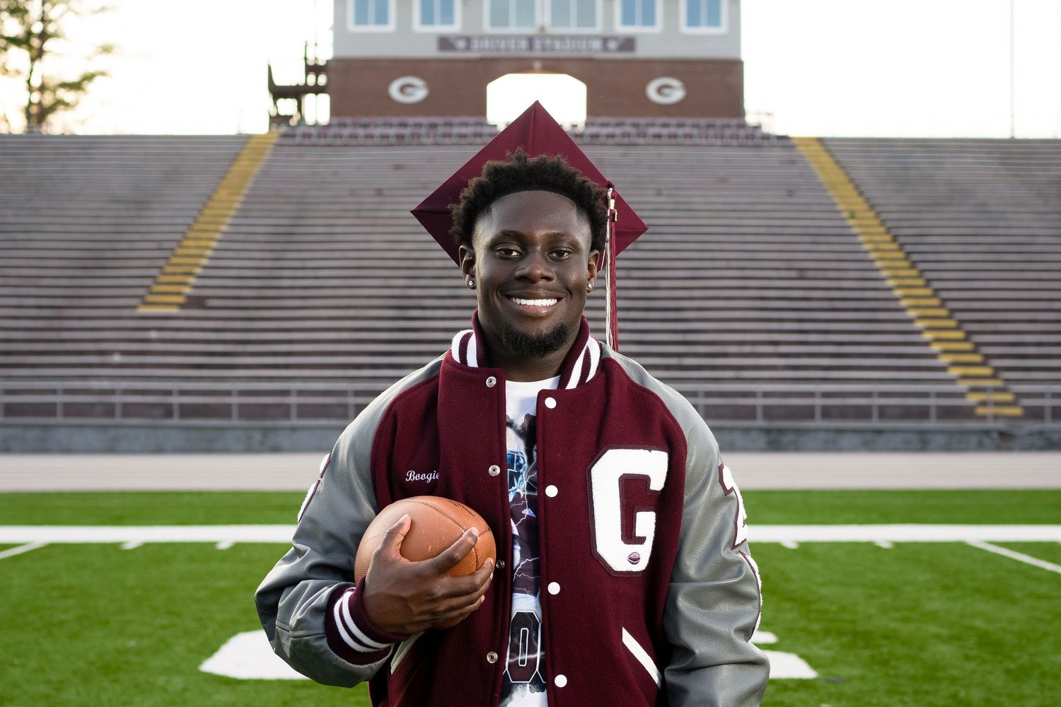 Graduate holding football while posing in varsity jacket at stadium.