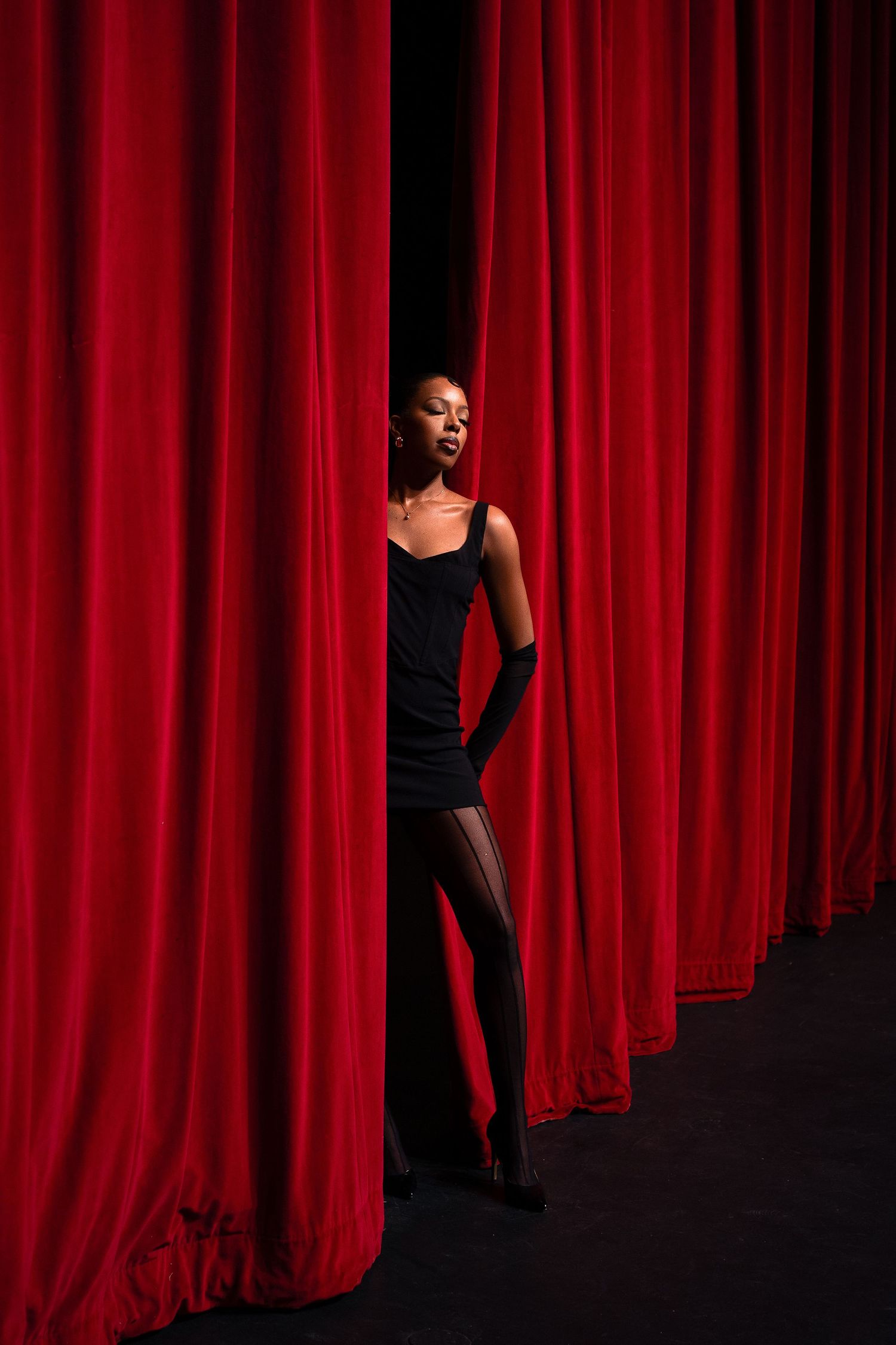 Dramatic figure in black outfit stands between luxurious red theater curtains in moody lighting.