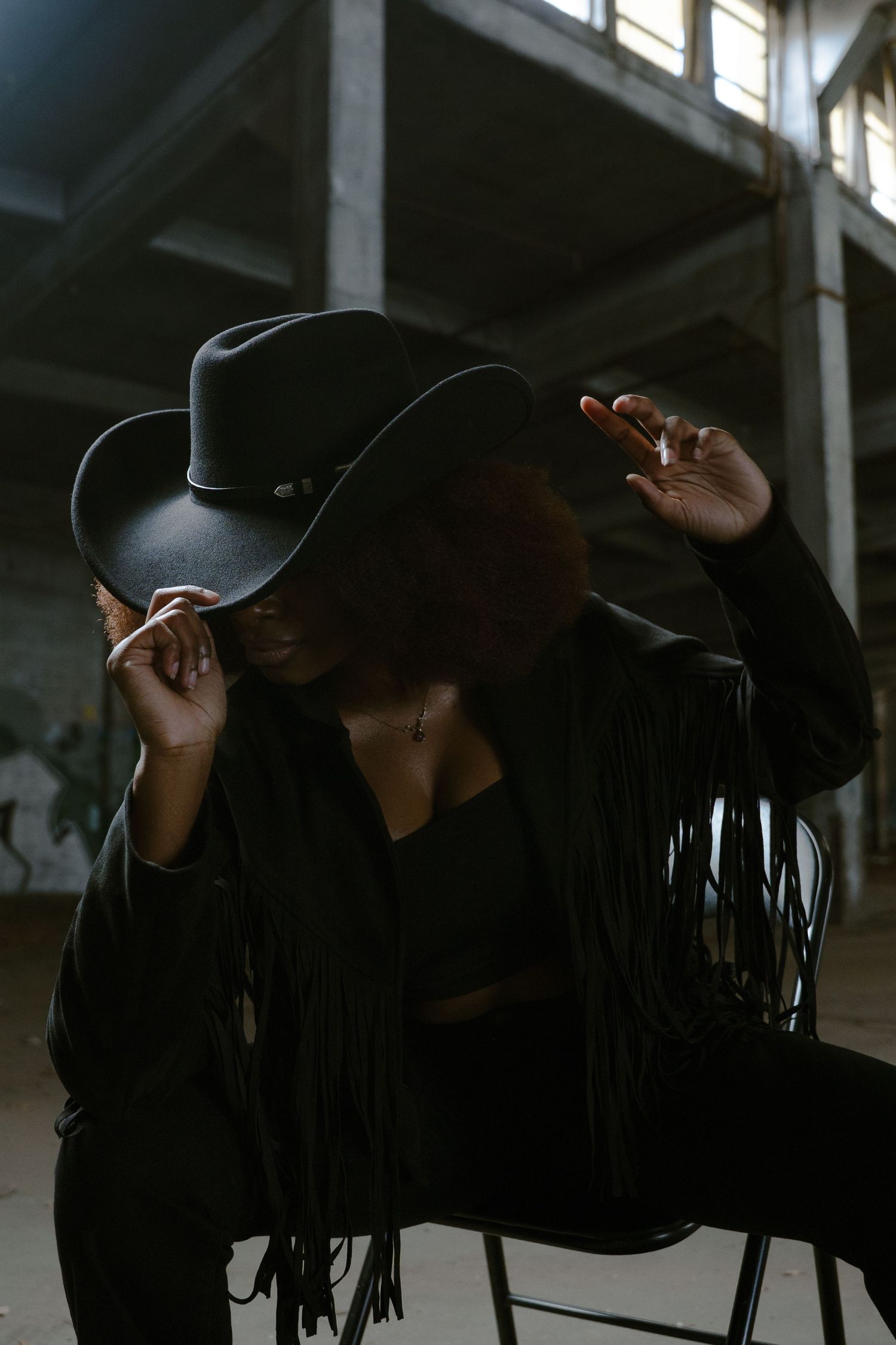 Stylish figure in black ensemble and wide-brimmed cowboy hat poses in industrial setting.