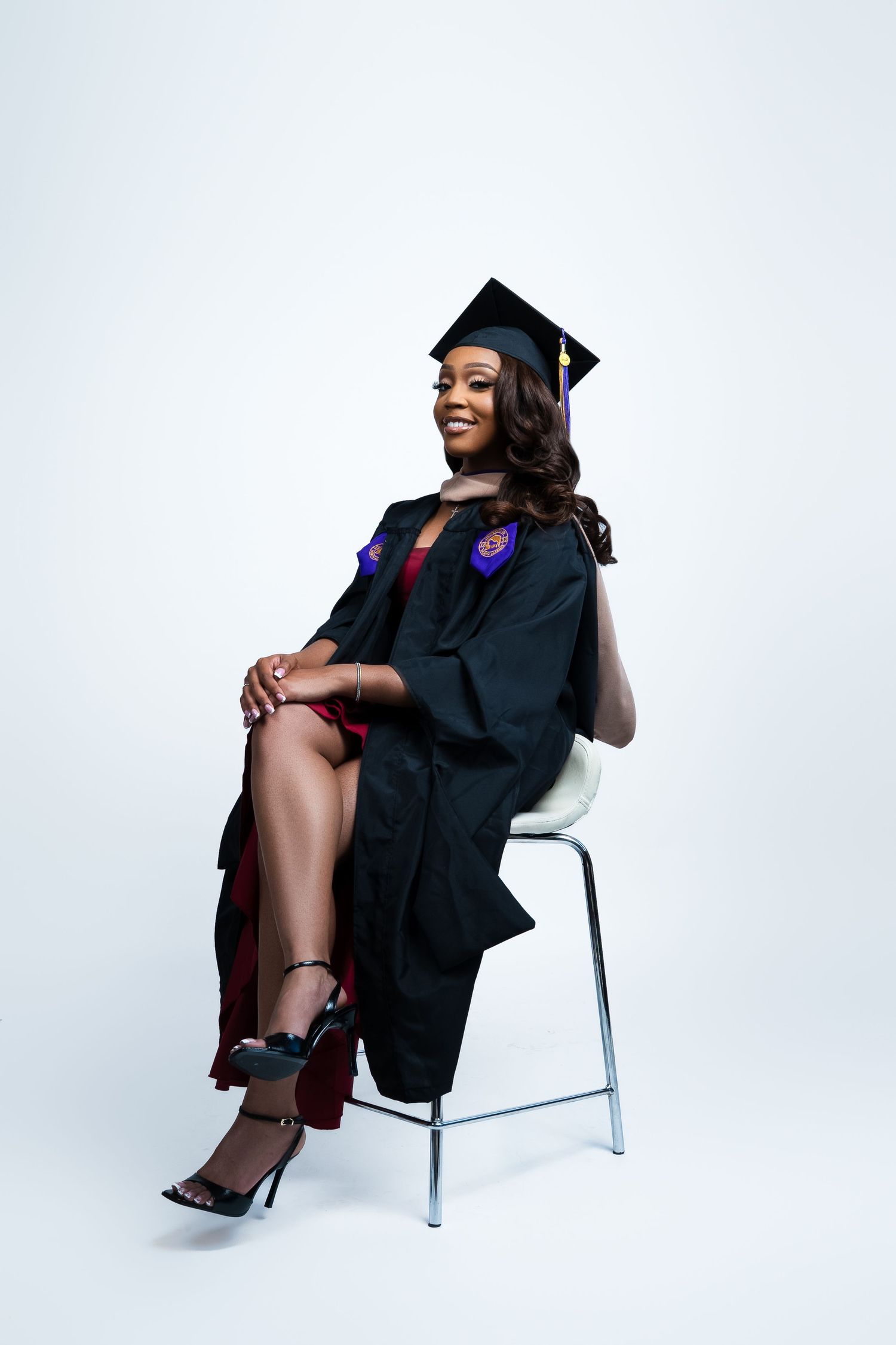 Graduate in traditional black cap and gown sits elegantly on white chair.