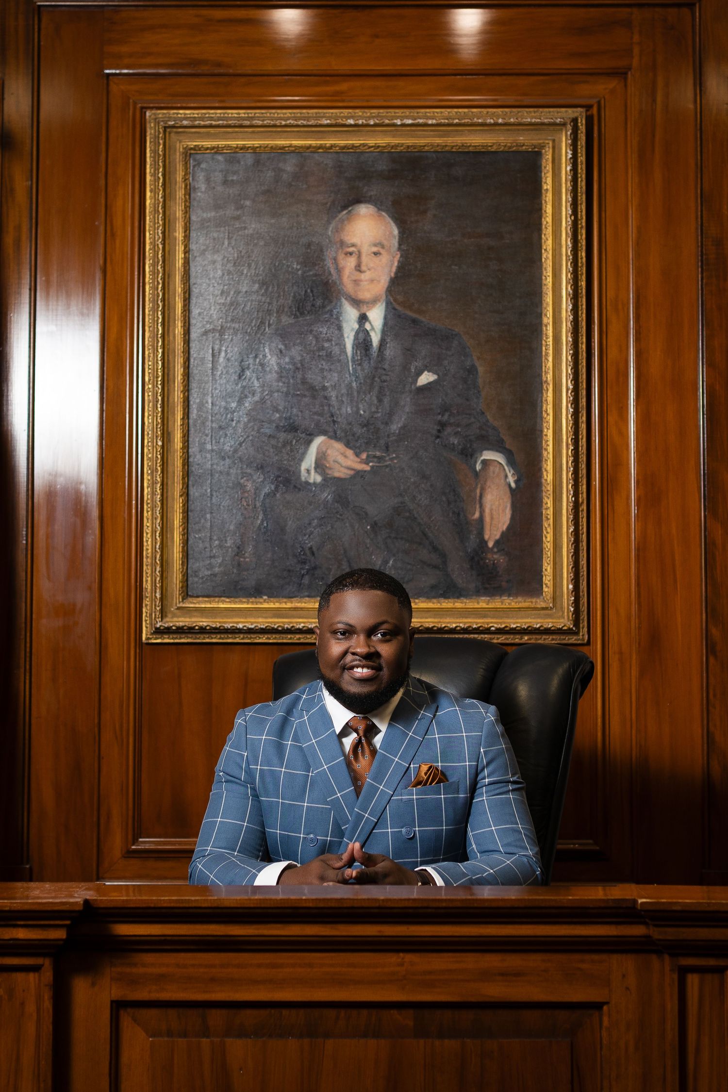 Professional seated at executive desk beneath formal oil portrait in wood-paneled office.
