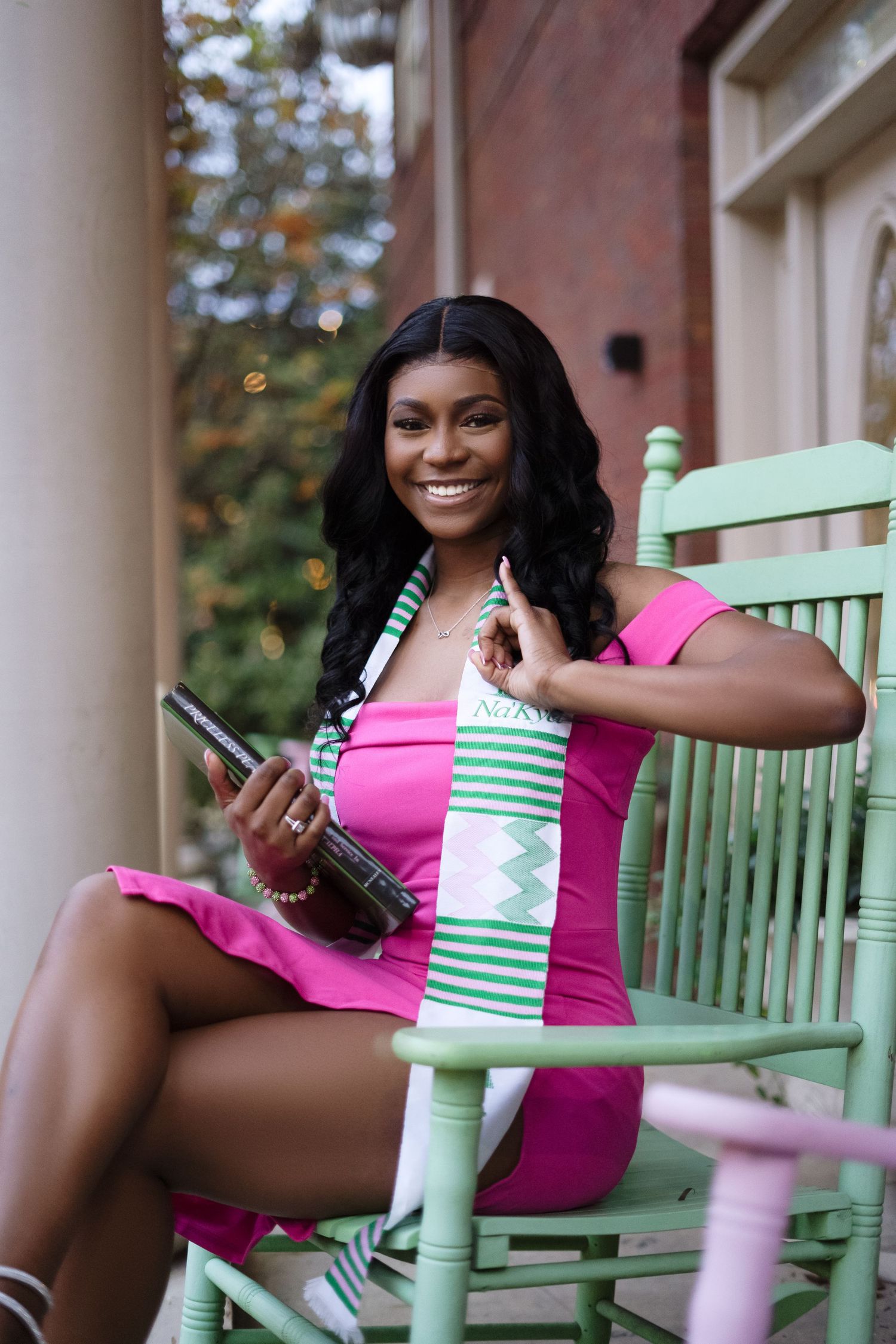 Graduate relaxes in pink dress on mint green rocking chair with graduation cap.