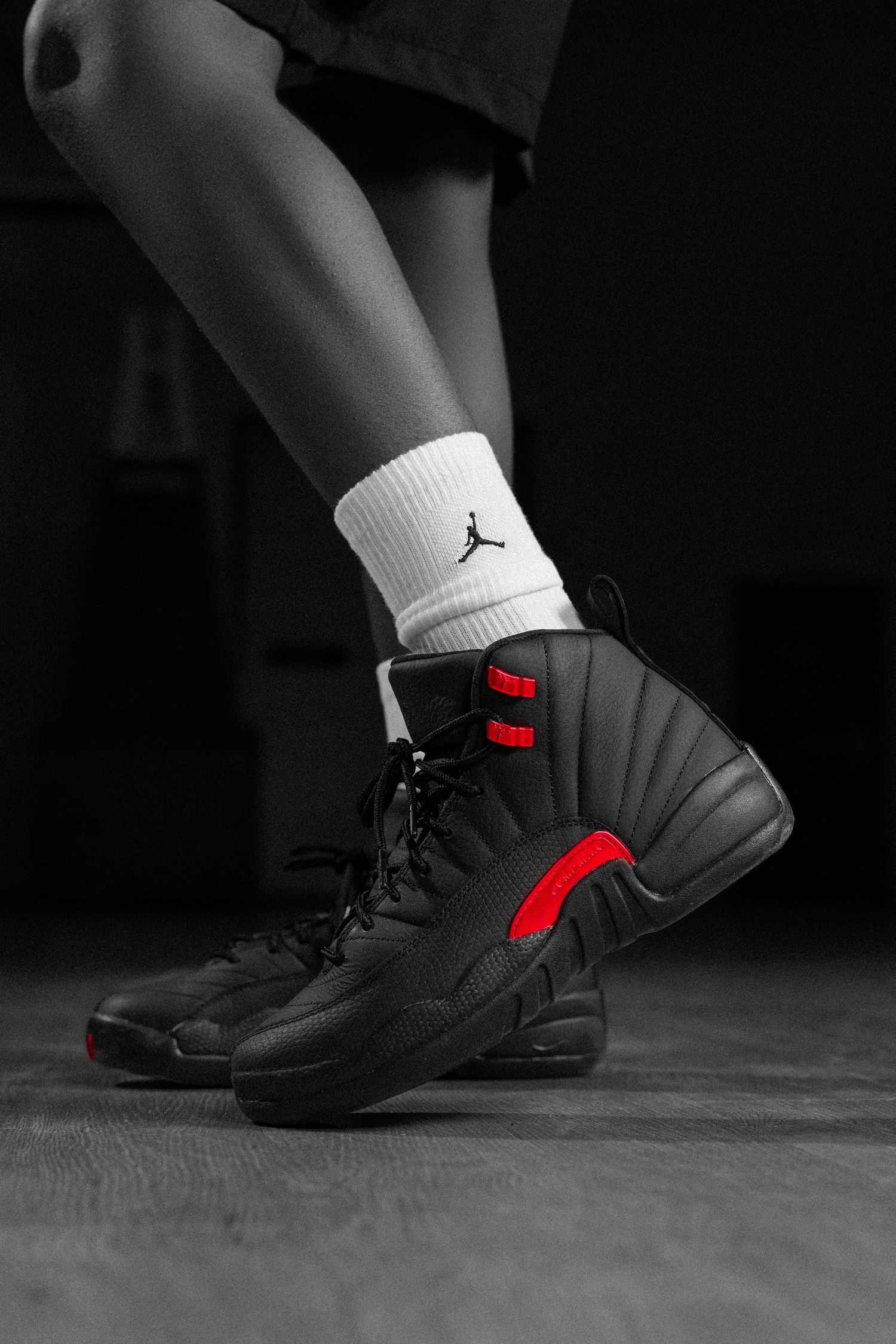 Black and red Air Jordan 12 sneakers worn with white Jordan socks, styled in a dark moody setting.