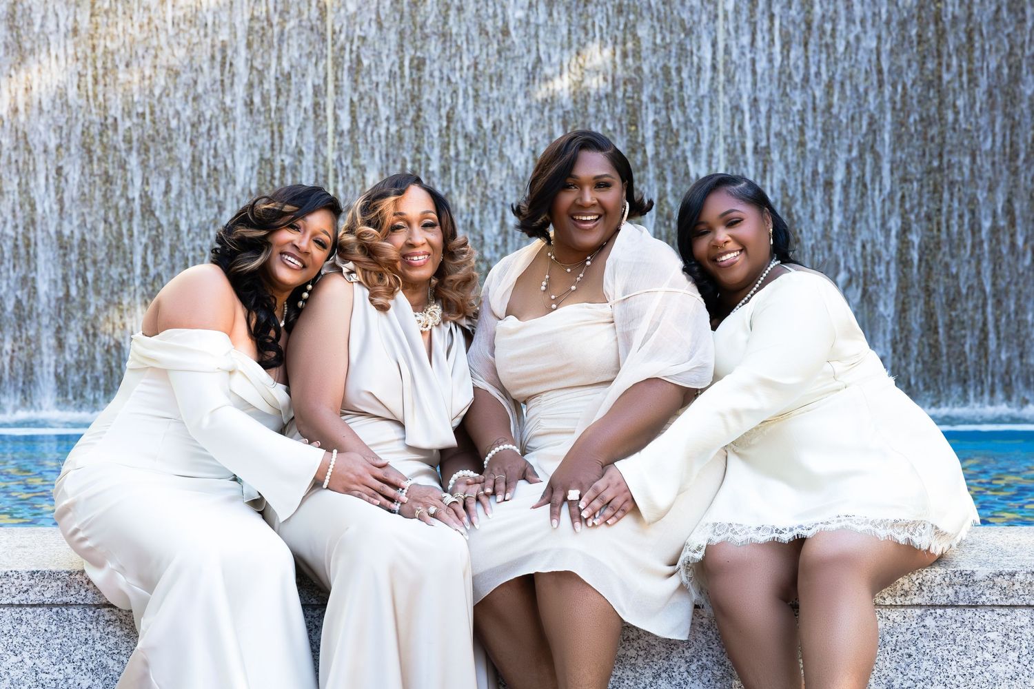 Four women in white outfits pose together smiling in front of a fountain.