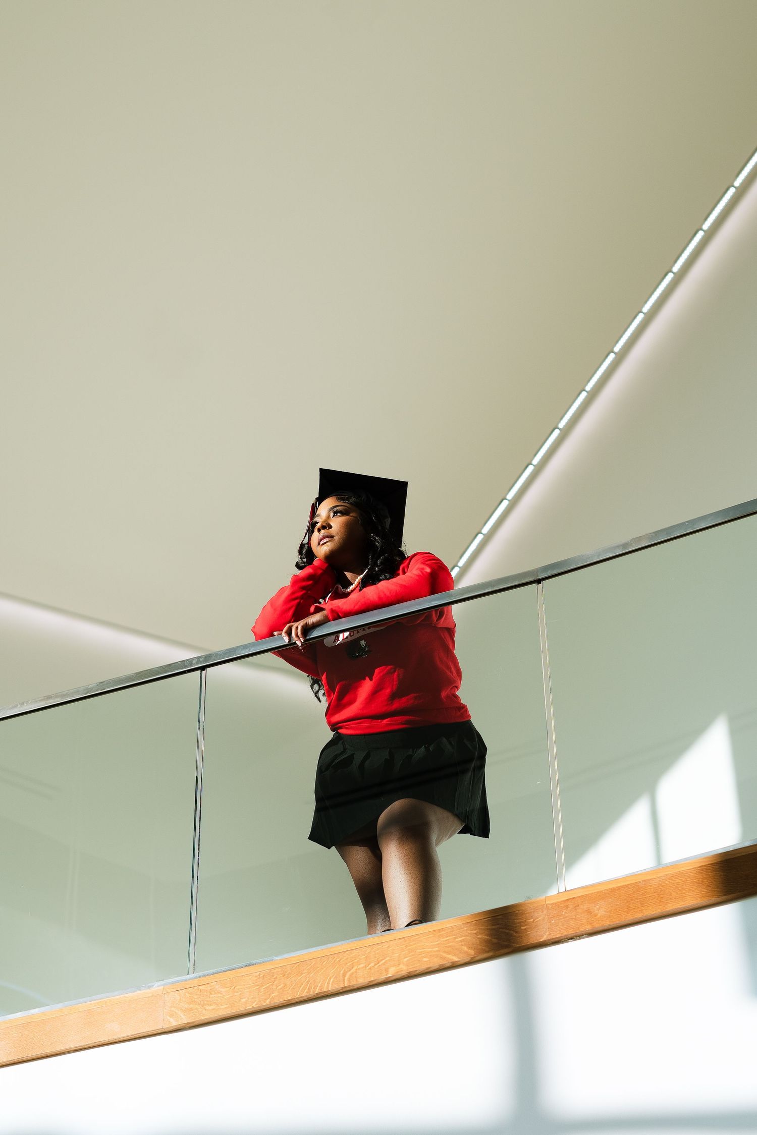 Graduate in red sweatshirt leans on railing in modern architecture setting with dramatic lighting.