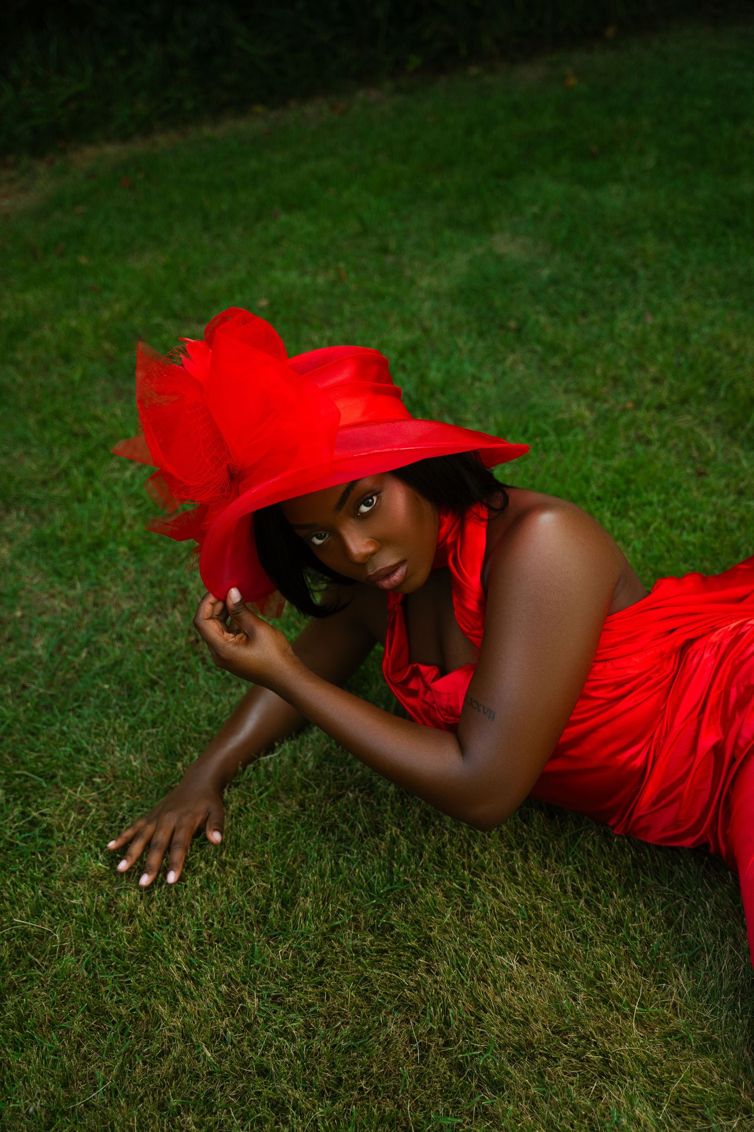 A person in a vibrant red dress and matching headpiece lies gracefully on lush green grass in an artistic pose.