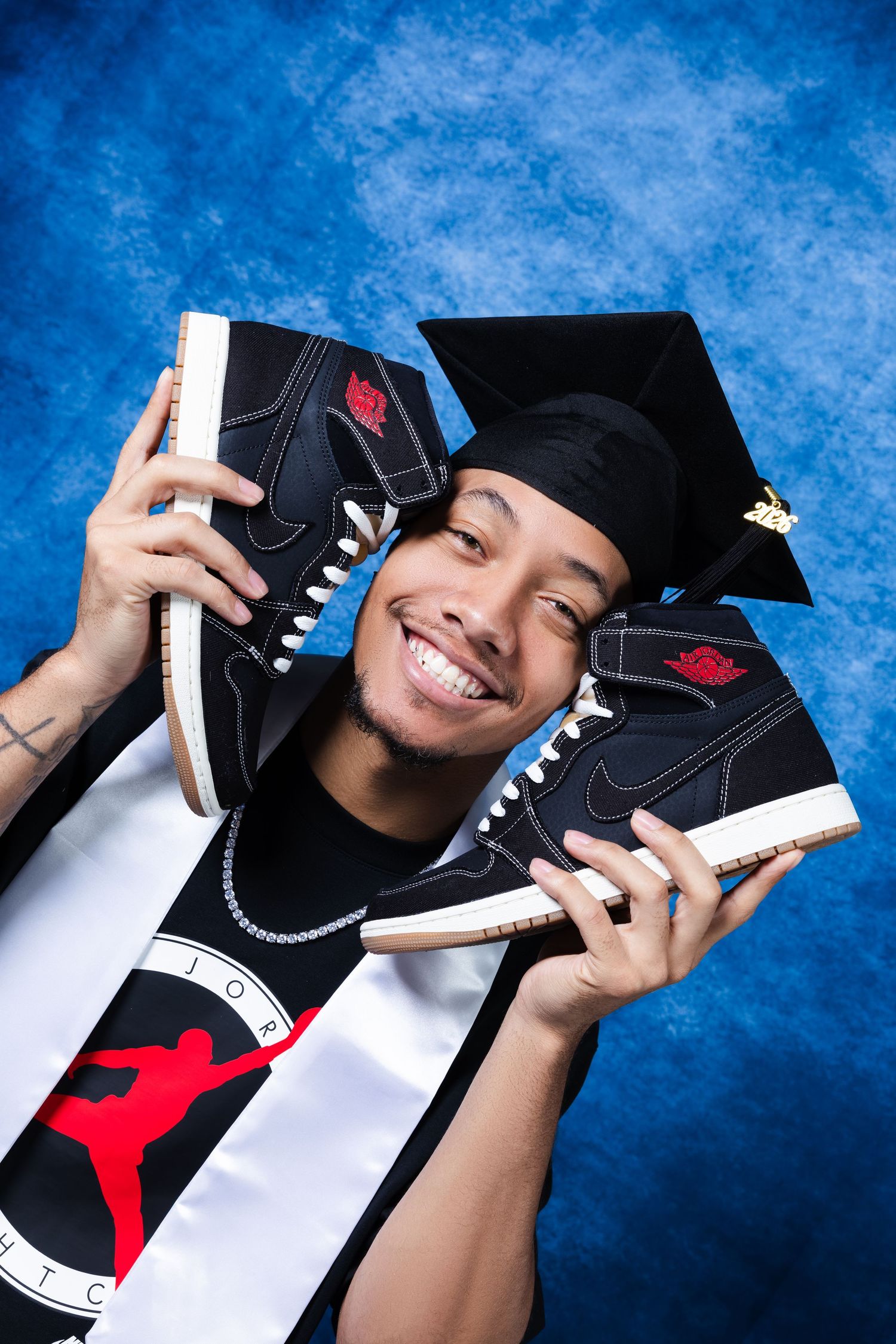 Smiling graduate holding Air Jordan 1 sneakers, wearing graduation cap and Jordan Brand sash against blue backdrop.