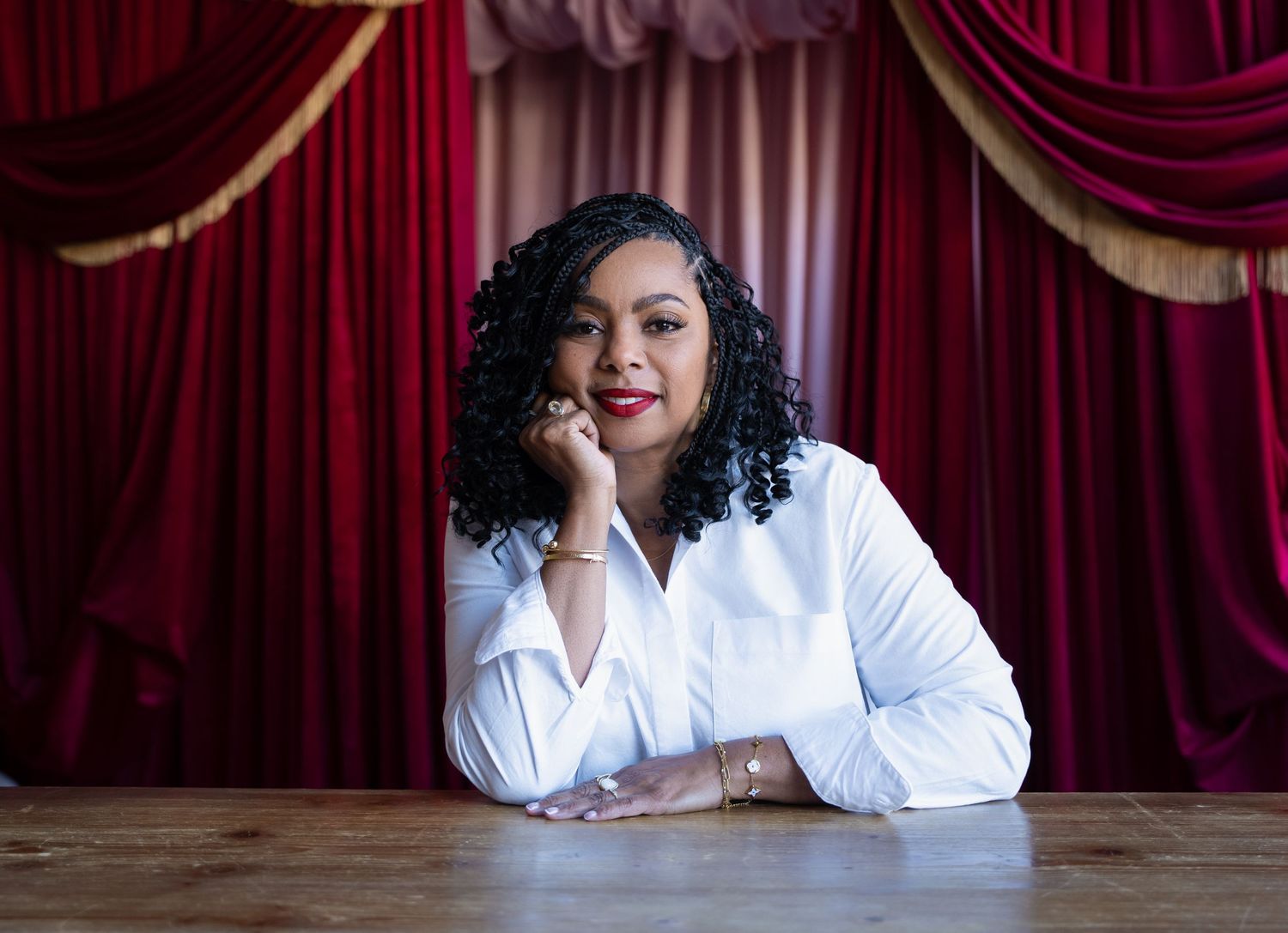 Woman in white blouse poses confidently at wooden table before elegant red velvet theater curtains.