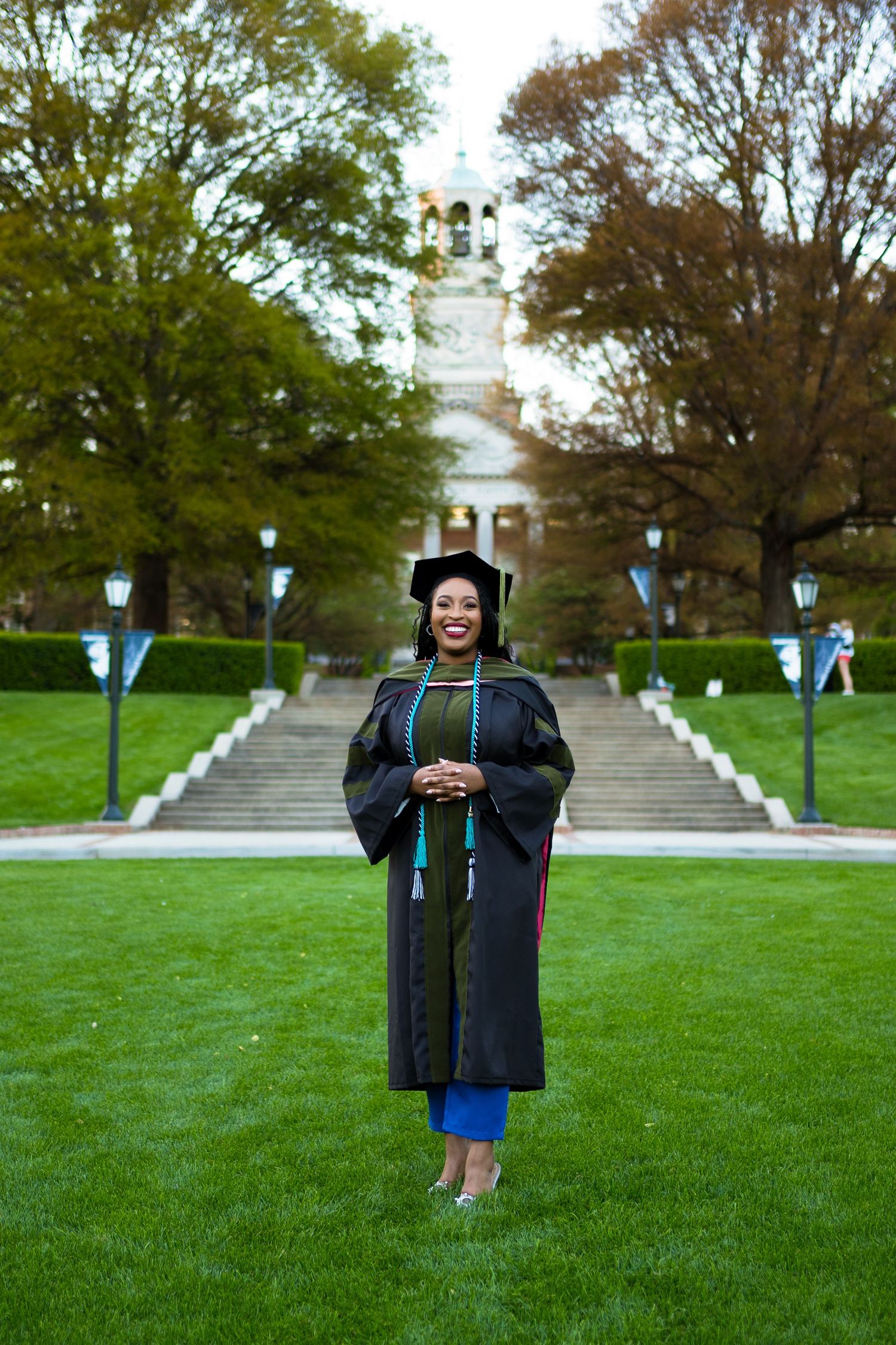 Graduate in traditional academic regalia stands on lawn path leading to classic university building.