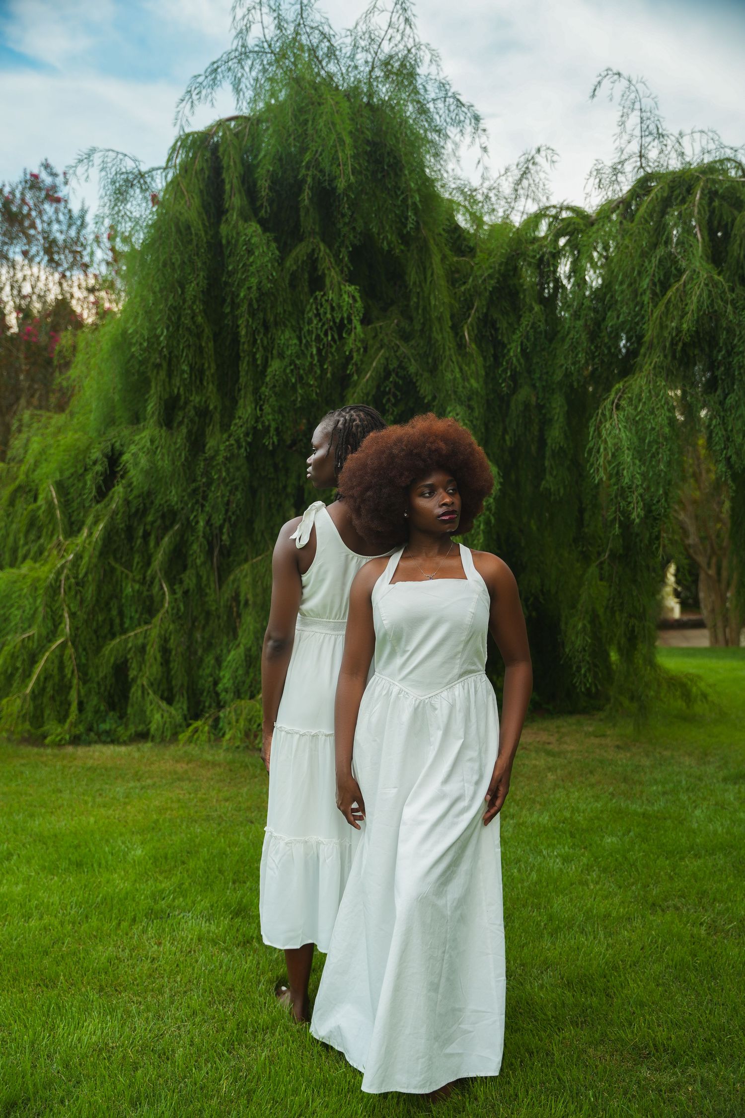 A figure in a flowing white dress poses gracefully against a backdrop of drooping willow trees in a lush green park setting.