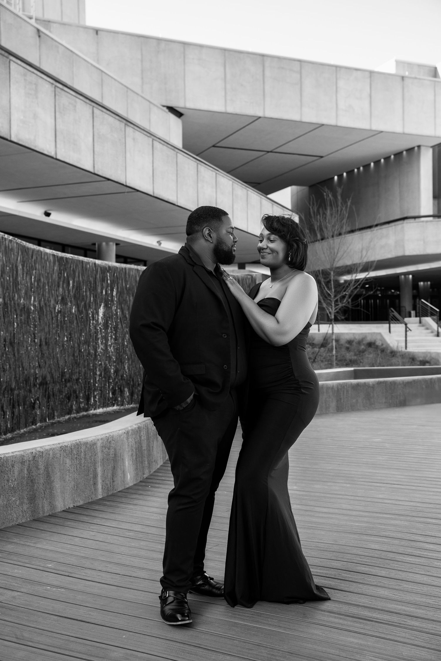Black and white photo of a couple in formal attire sharing a romantic moment outside a modern building.