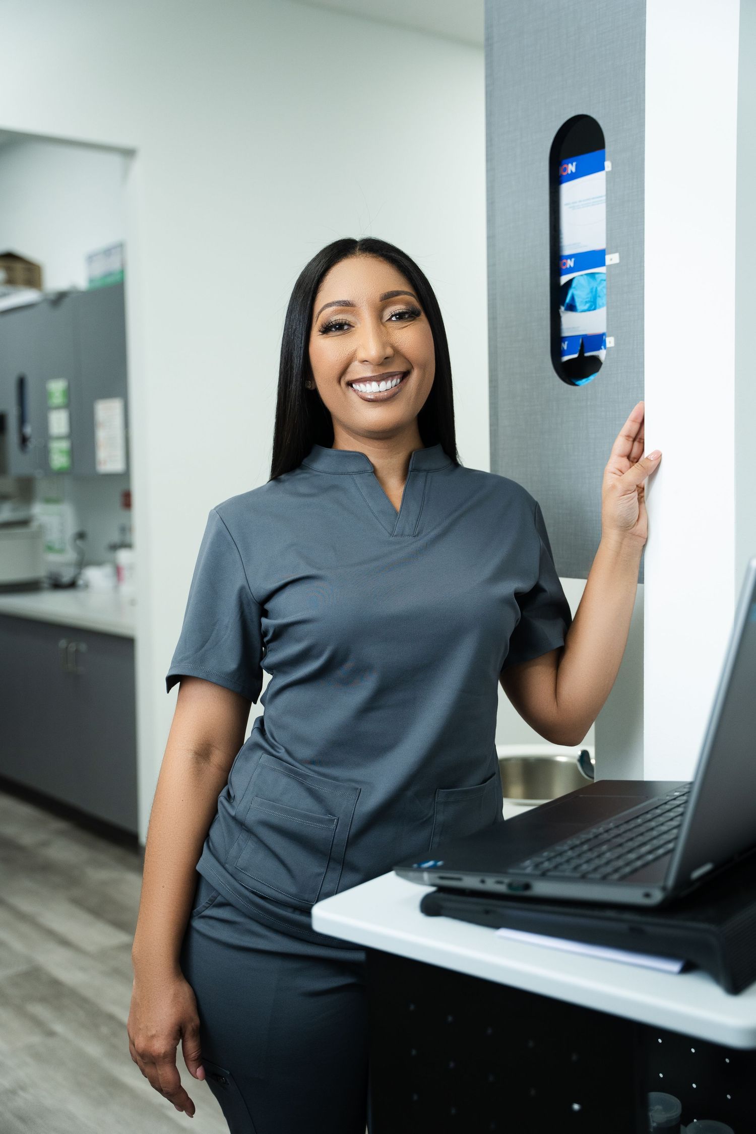 Smiling female healthcare professional in gray scrubs standing at a medical office workstation with laptop.