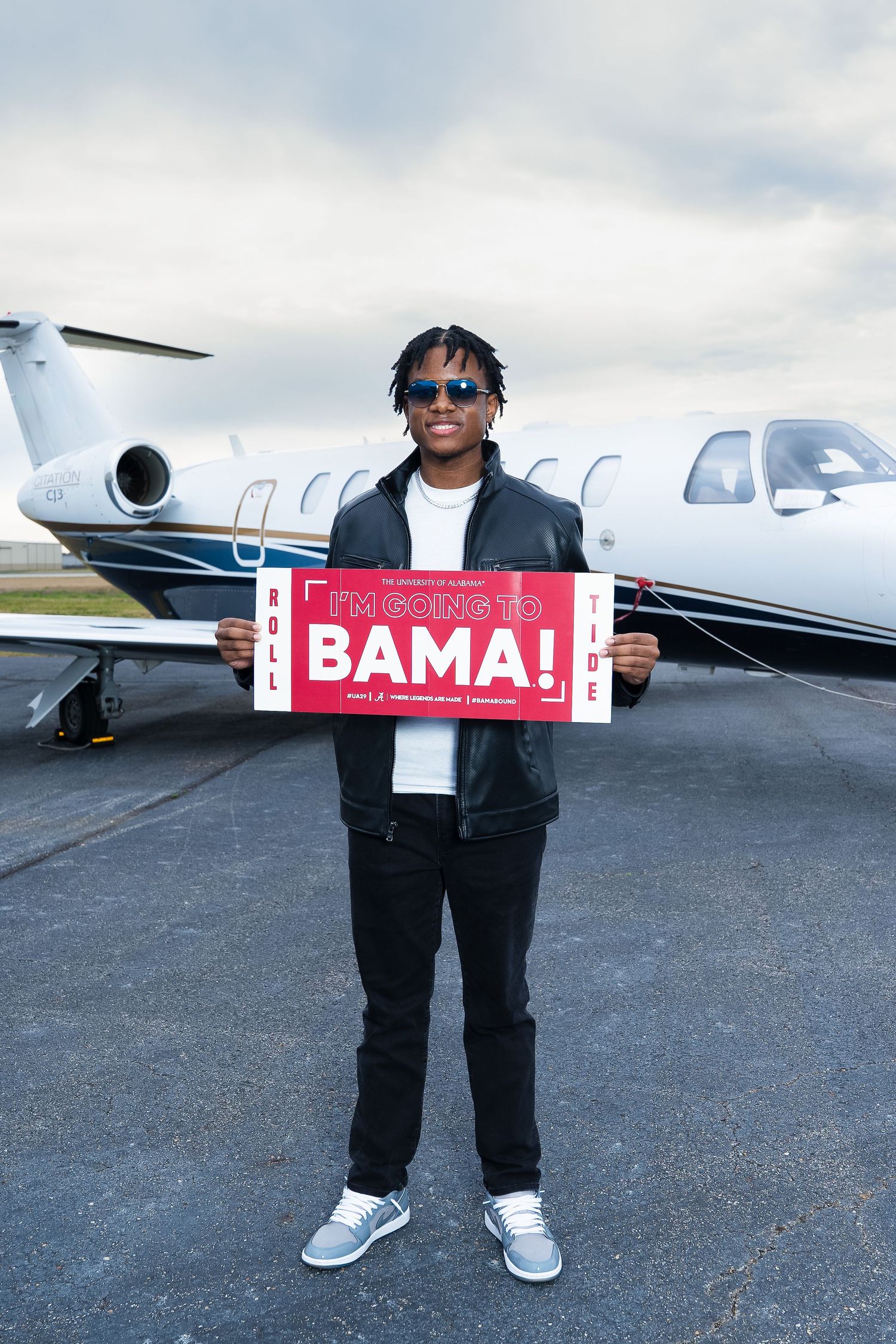 Person holding a 'Roll Bama!' sign while standing in front of a private jet.