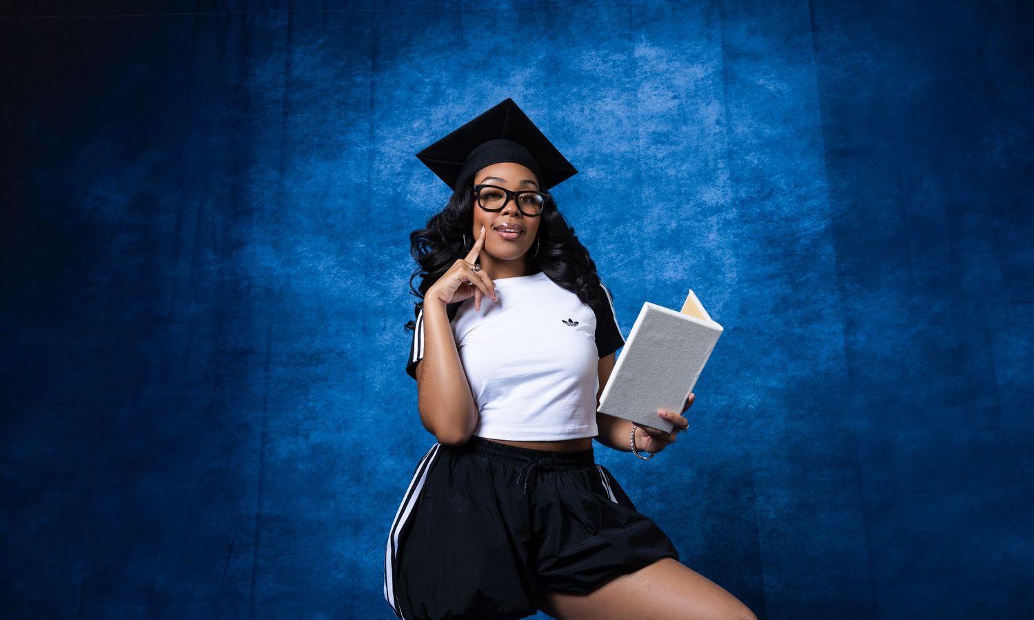 A young woman in graduation cap and glasses poses thoughtfully with a book against a blue background.
