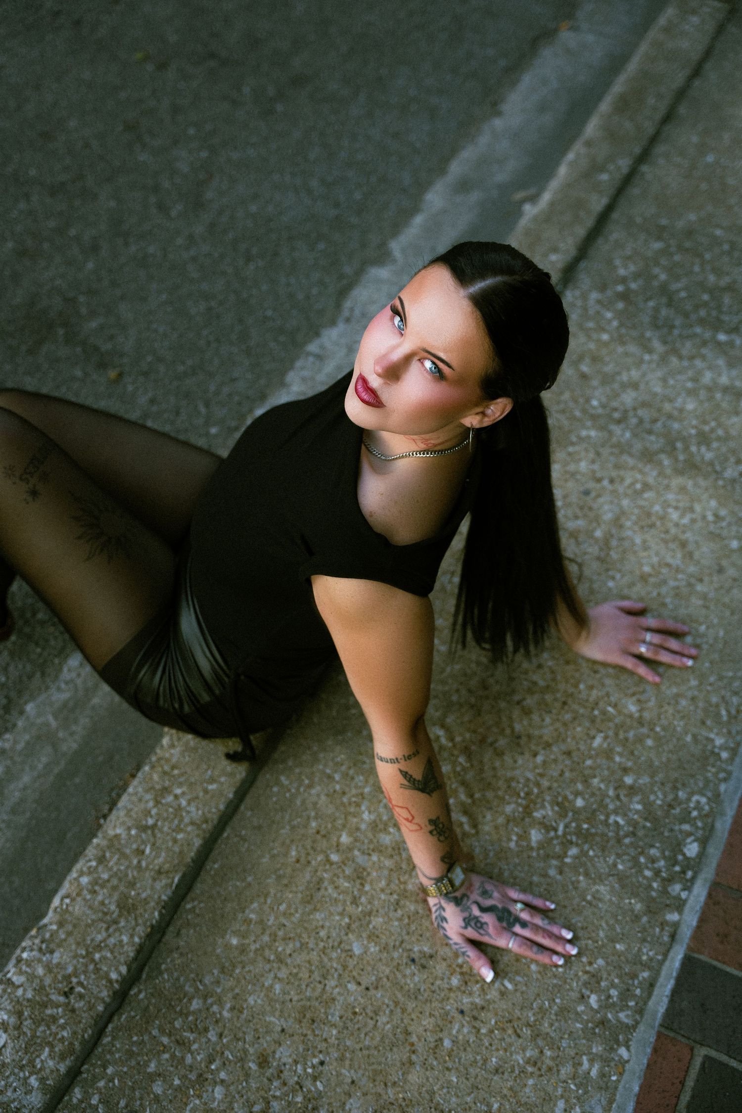 Woman with dark hair and red lips in black outfit poses on concrete steps, showcasing tattoos on her hand and arm.