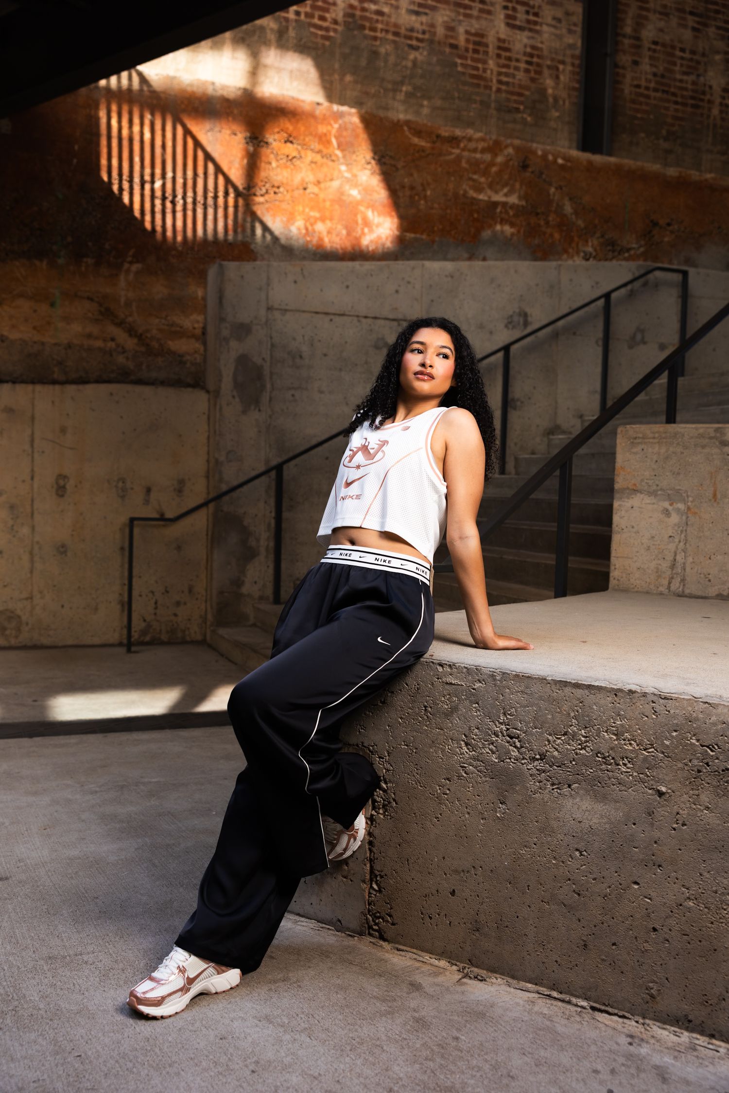A person in casual streetwear poses against a concrete wall with dramatic lighting and shadows in an industrial space.