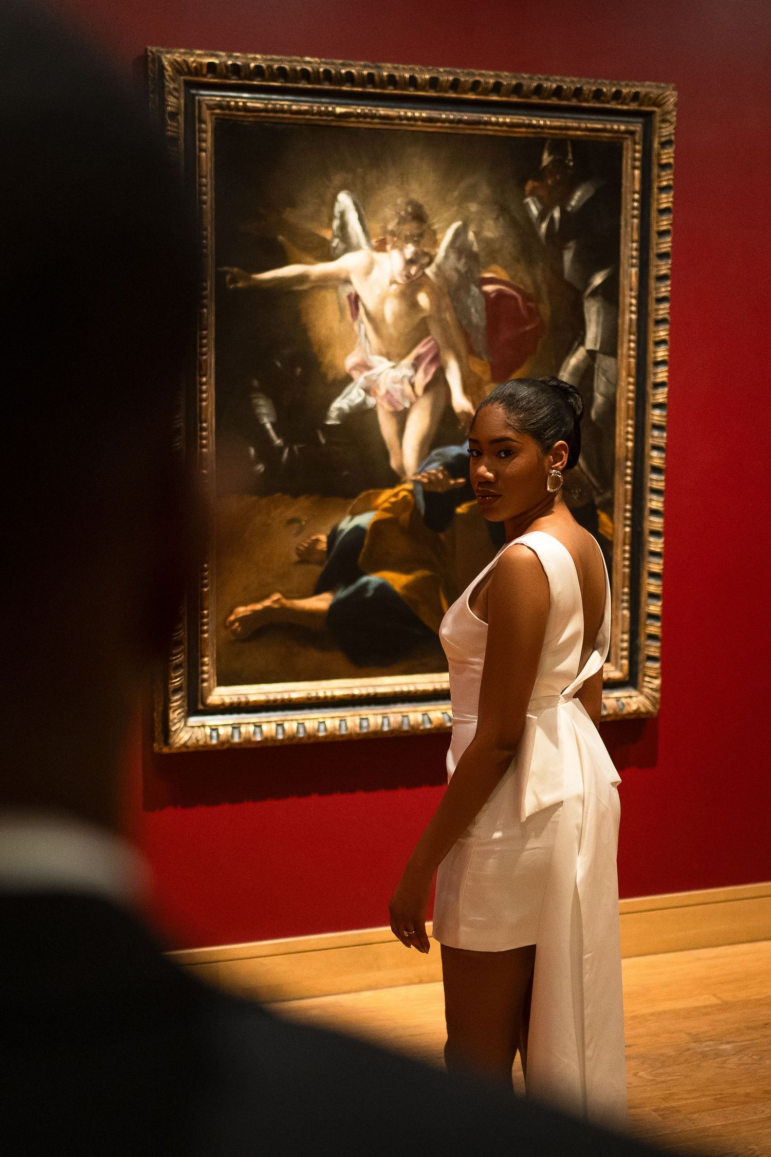 Woman in white dress views classical painting in art gallery with dramatic lighting.