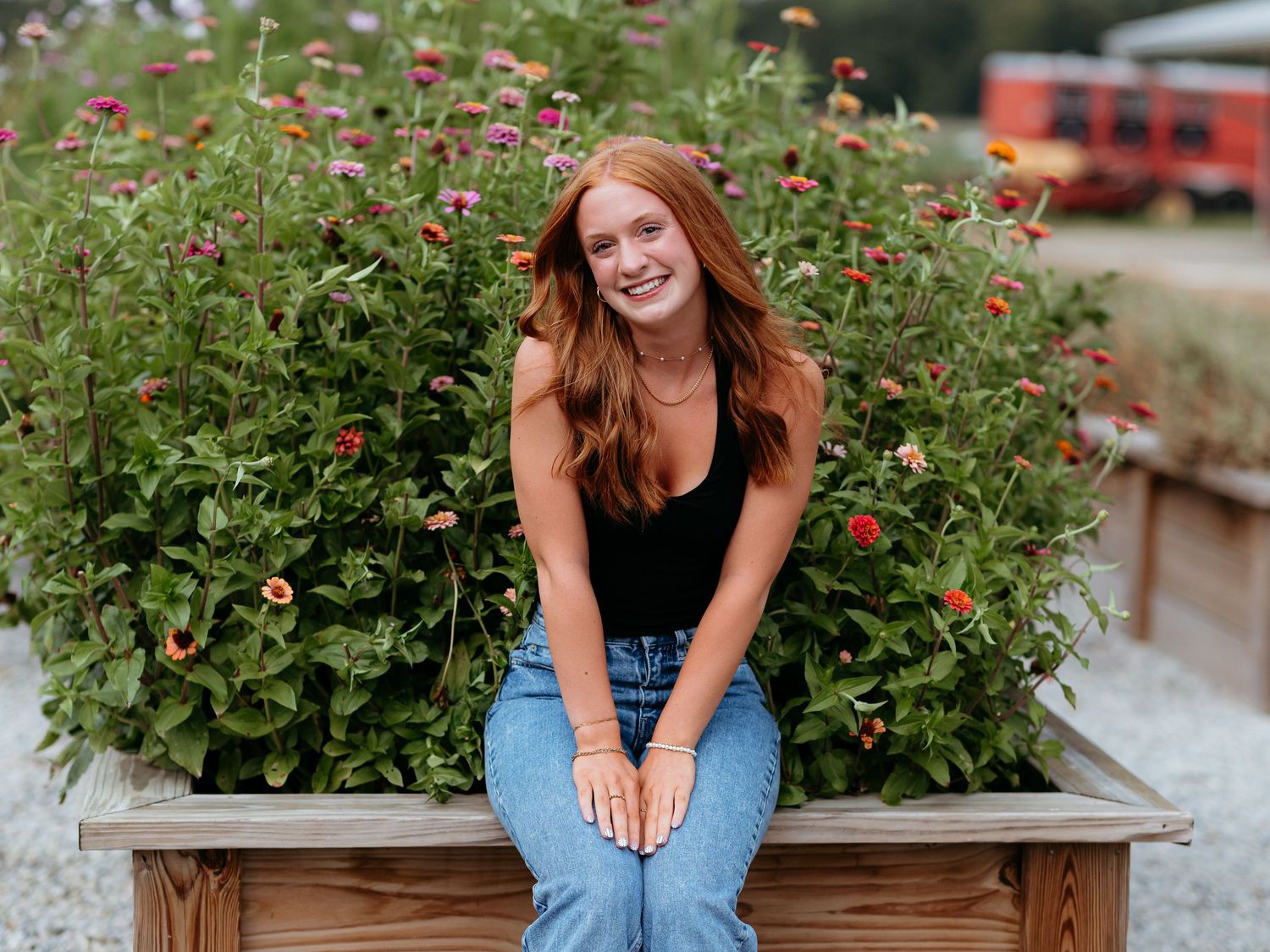 A senior girl sits on a wooden planter box surrounded by colorful flowering plants at heirloom grove venue. Senior photography sessions.