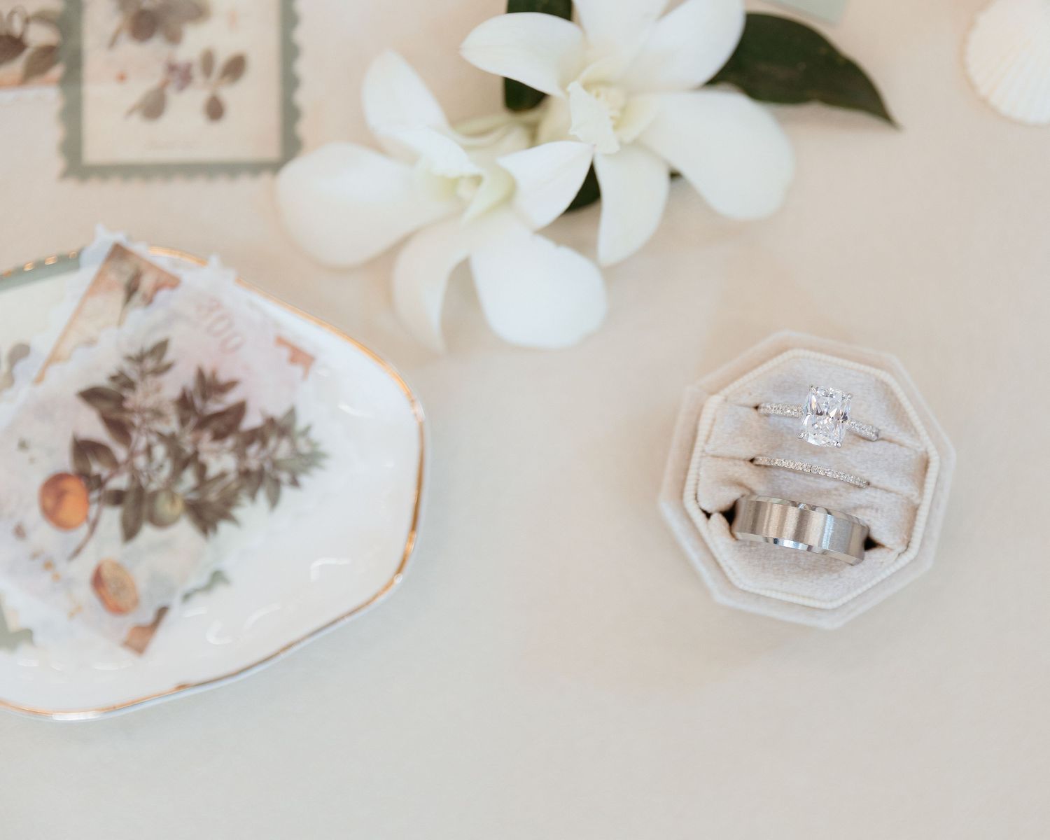 Wedding rings in a white box next to a white flower and decorative plate. Elegant wedding detail photographer. Bright and Airy photographer