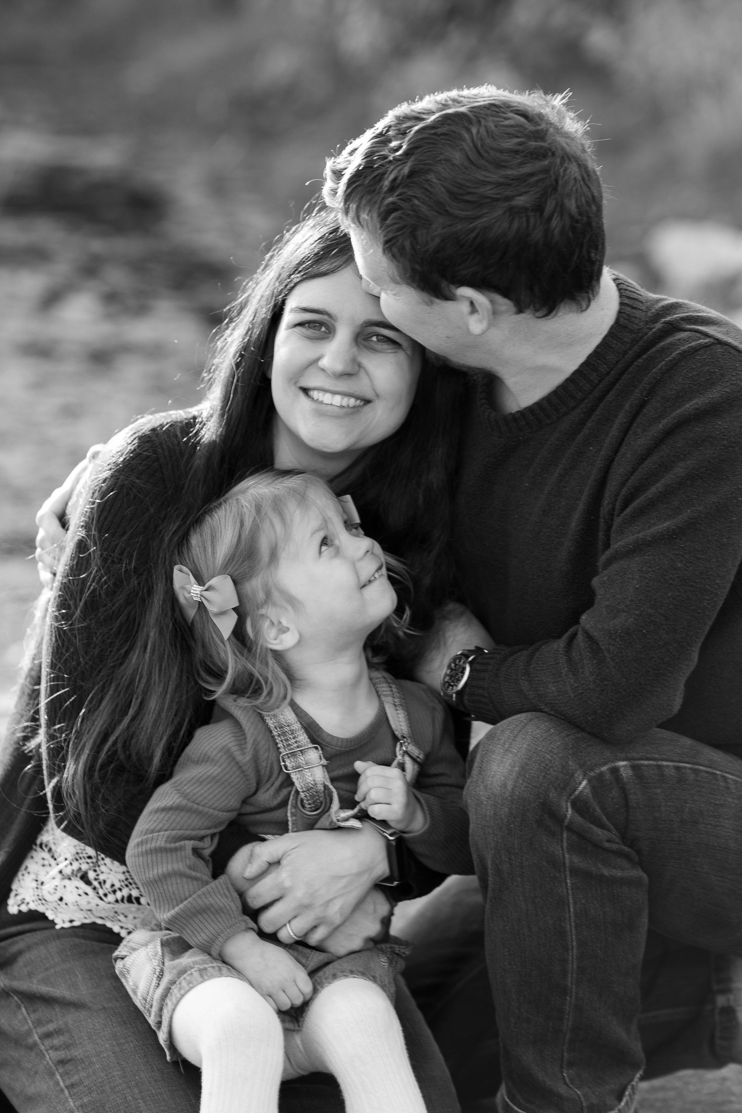 Black and white portrait of a loving family moment with parents and young child sitting together outdoors.