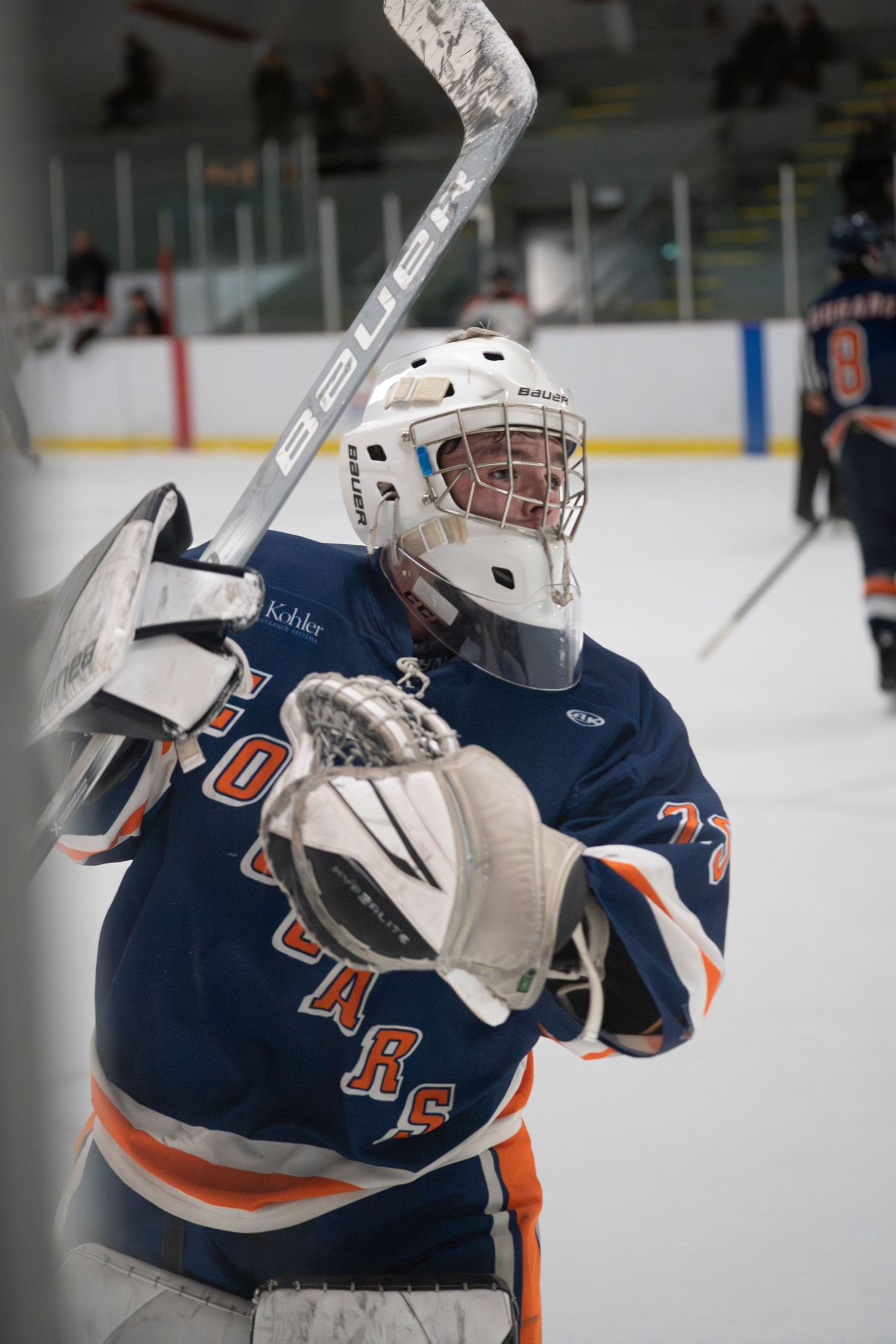 A hockey player in a navy and orange jersey celebrates with their stick raised on the ice during a game.