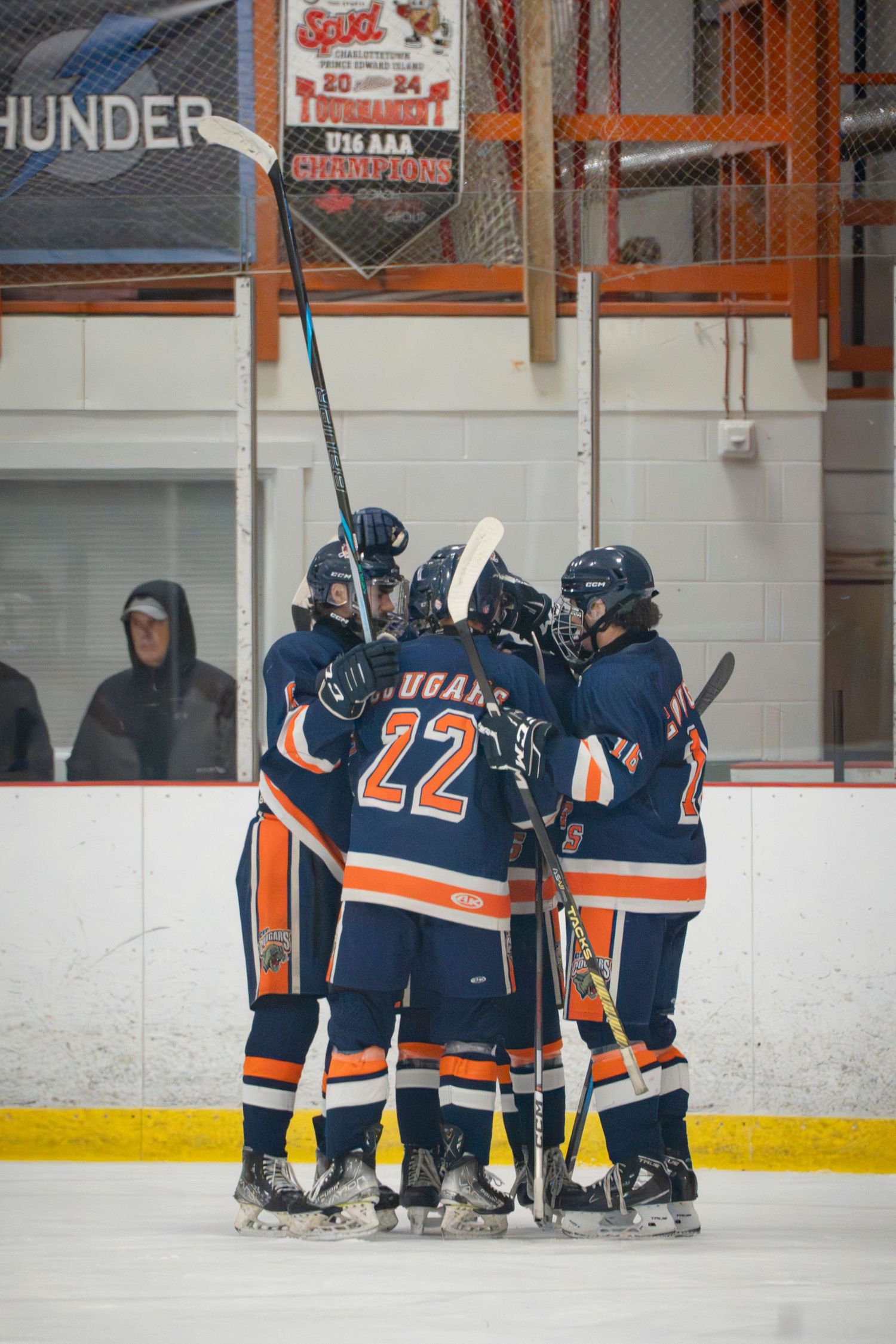 Youth hockey team huddles together on ice rink wearing navy blue and orange uniforms during game break.