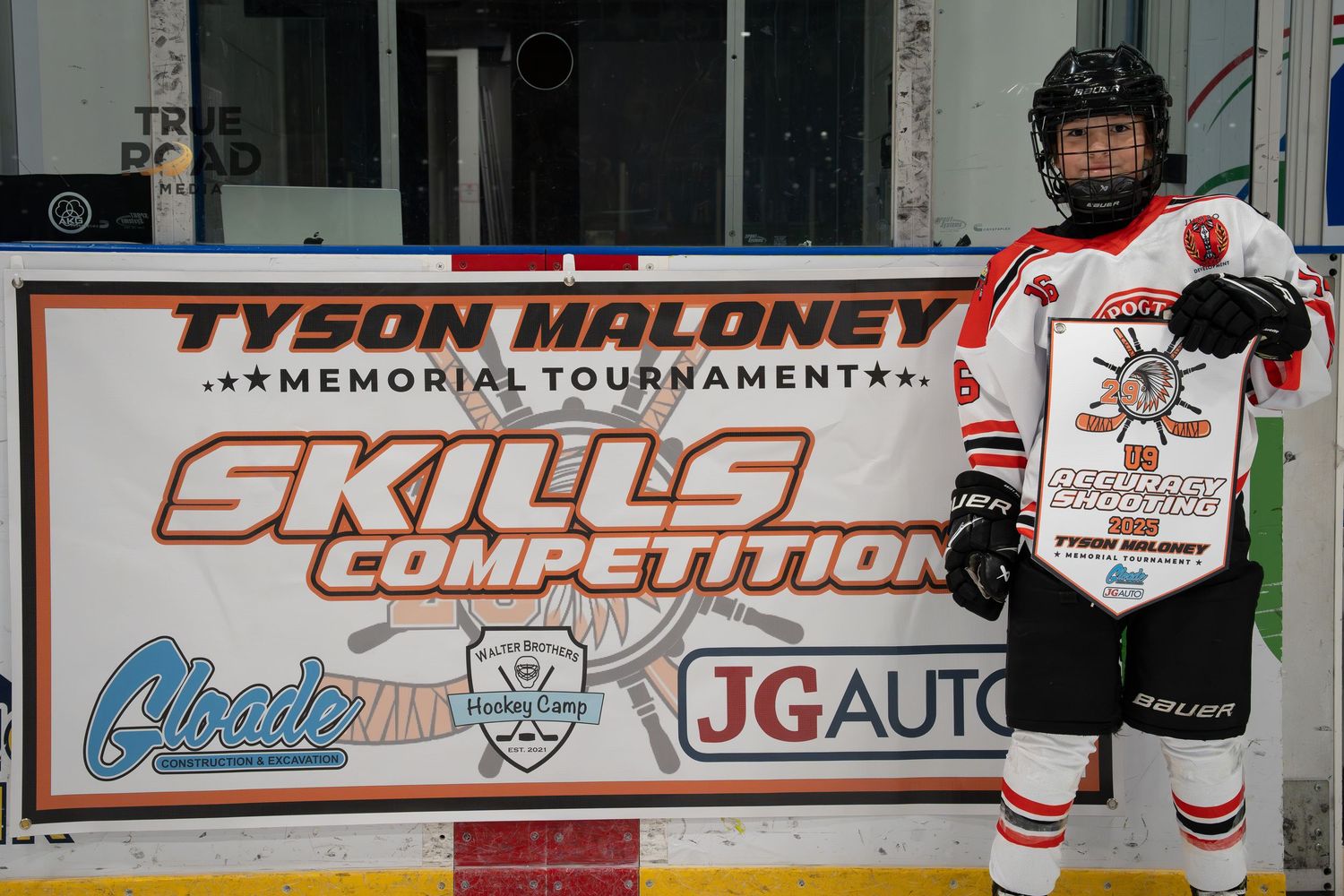 Hockey players line up along the boards during a game with advertisements and tournament signage visible in the background.