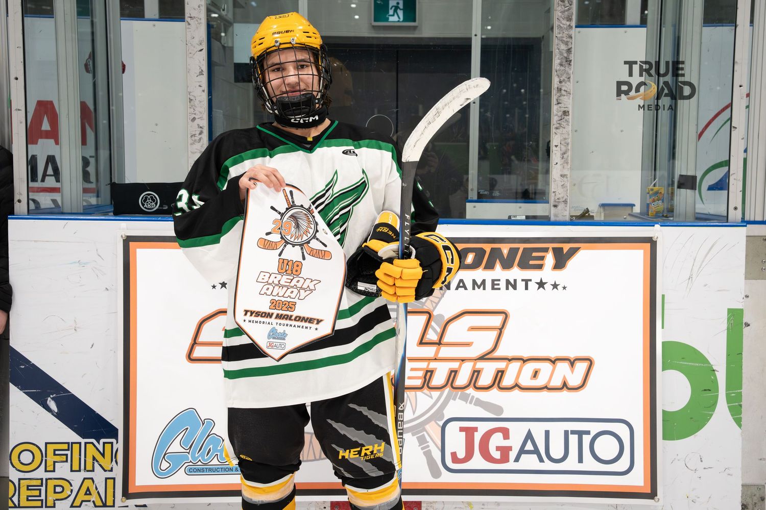 Hockey player in white and green jersey with yellow helmet stands near rink boards during game.