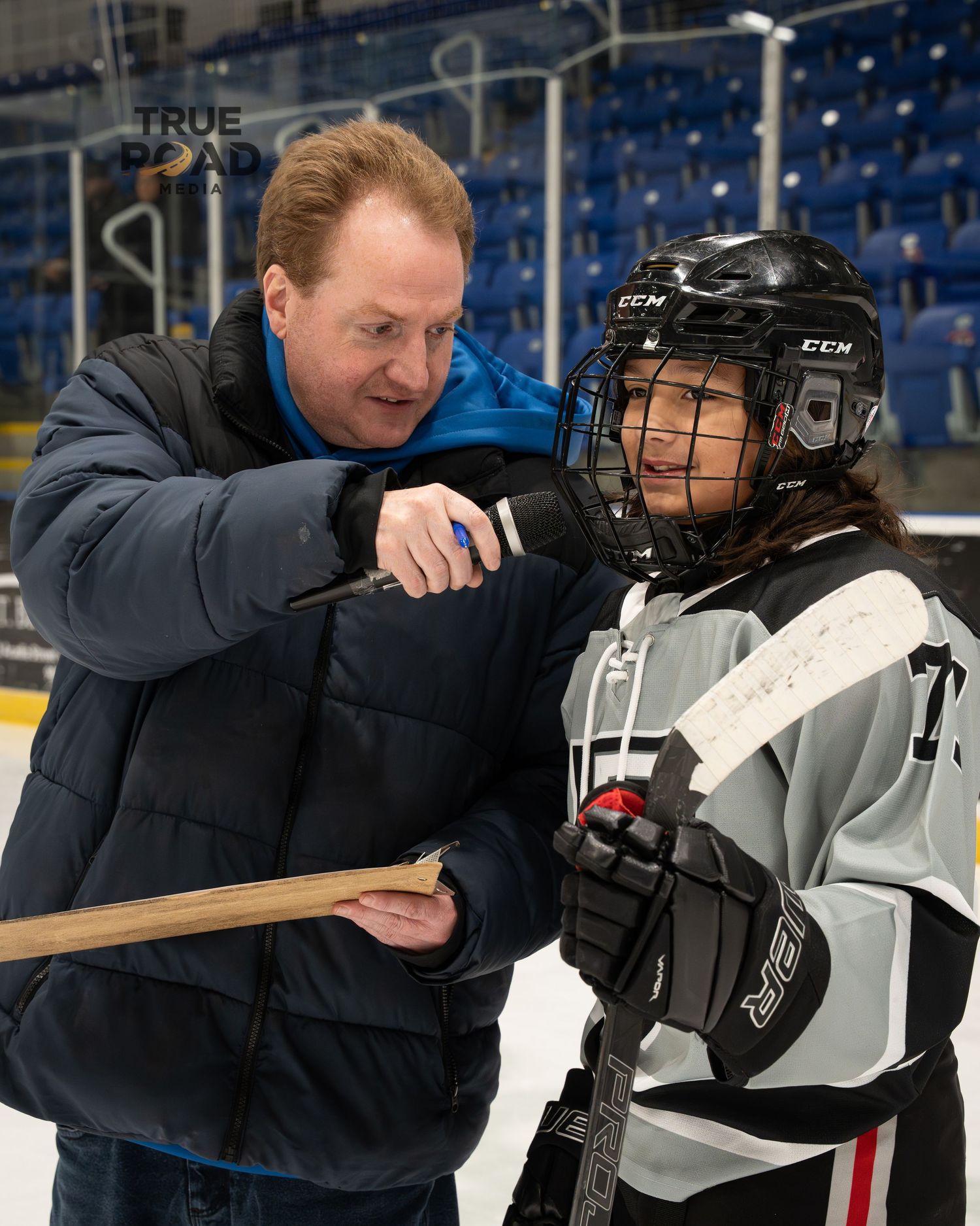 A hockey coach adjusts equipment on a young player wearing protective gear during practice at an indoor ice rink.