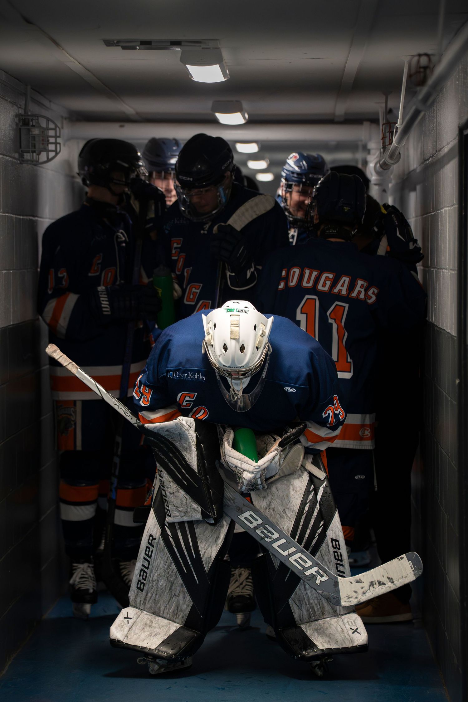 Hockey goalie equipment and jerseys line the walls of a dimly lit locker room with dark blue uniforms visible.