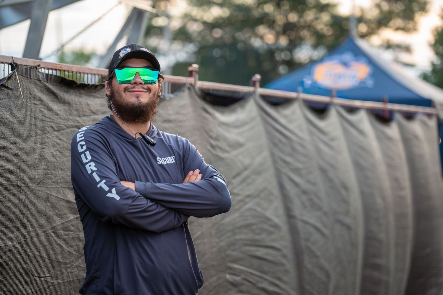 A person wearing a blue jacket and sunglasses smiles while standing in front of gray tents at an outdoor event.
