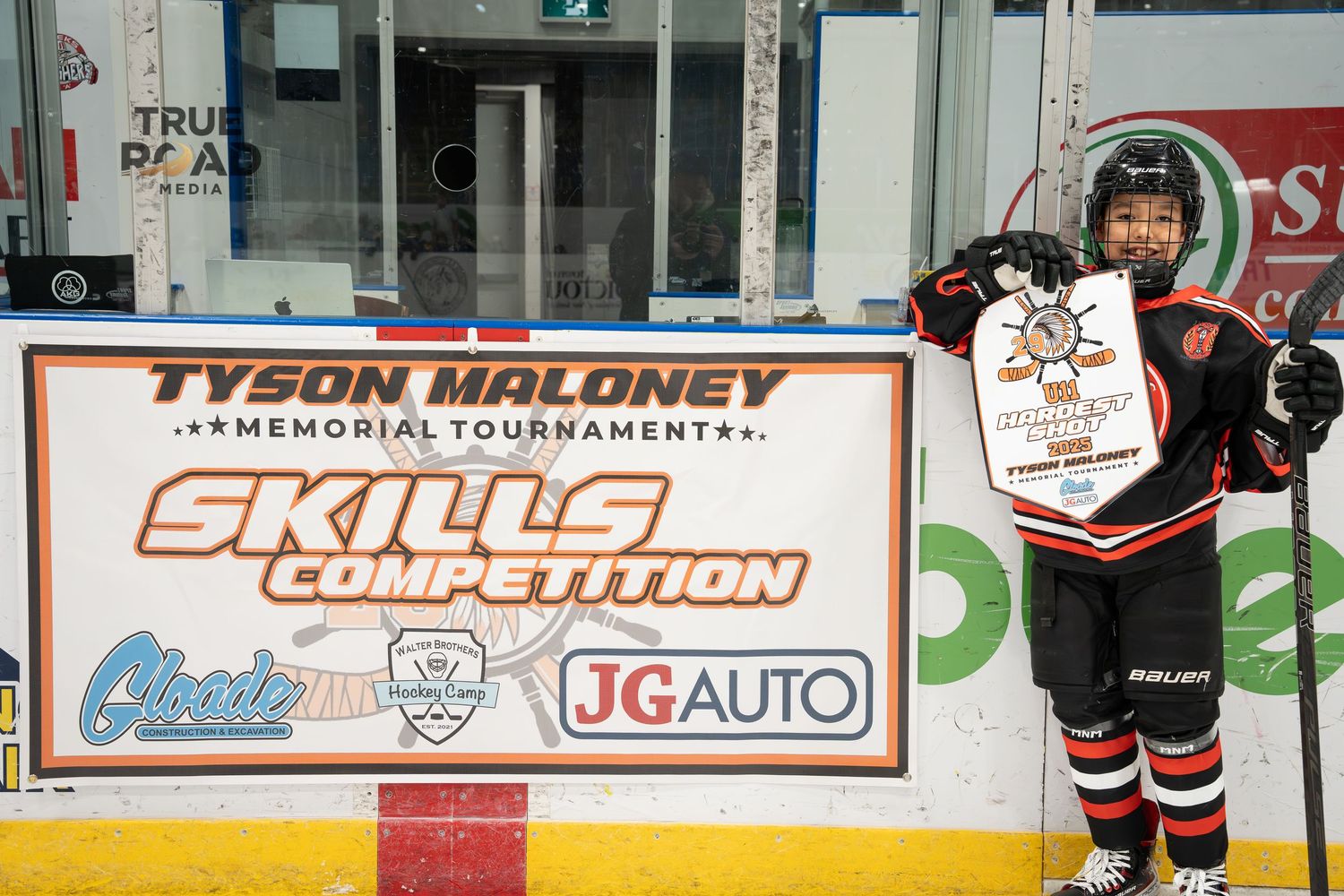 Hockey players sit in penalty box behind glass with advertising banners and sponsors visible in arena.