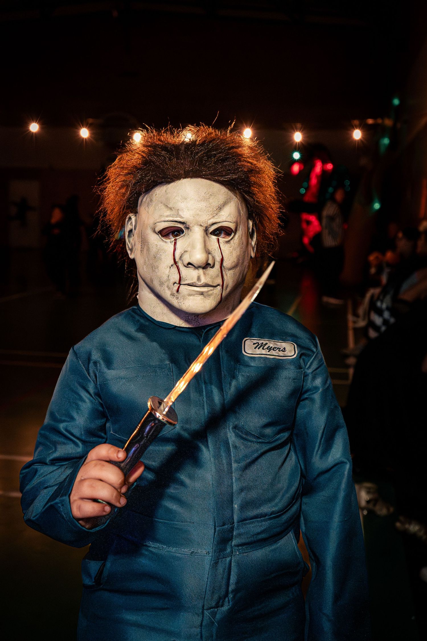 Person in Michael Myers Halloween costume with mask and blue coveralls holding knife in dark setting.