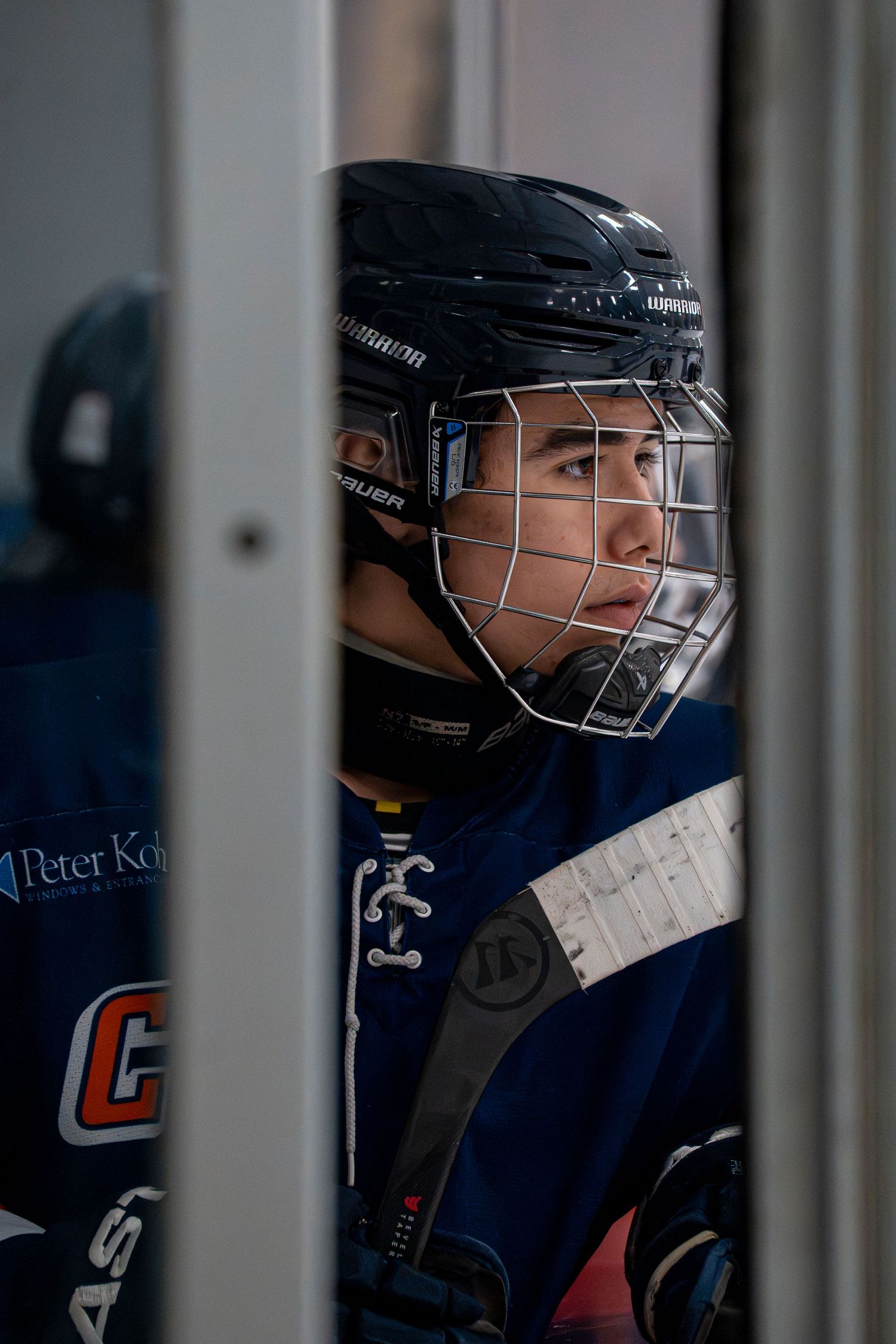 Hockey player wearing a helmet with face cage looks through the bars of a bench or barrier during a game.