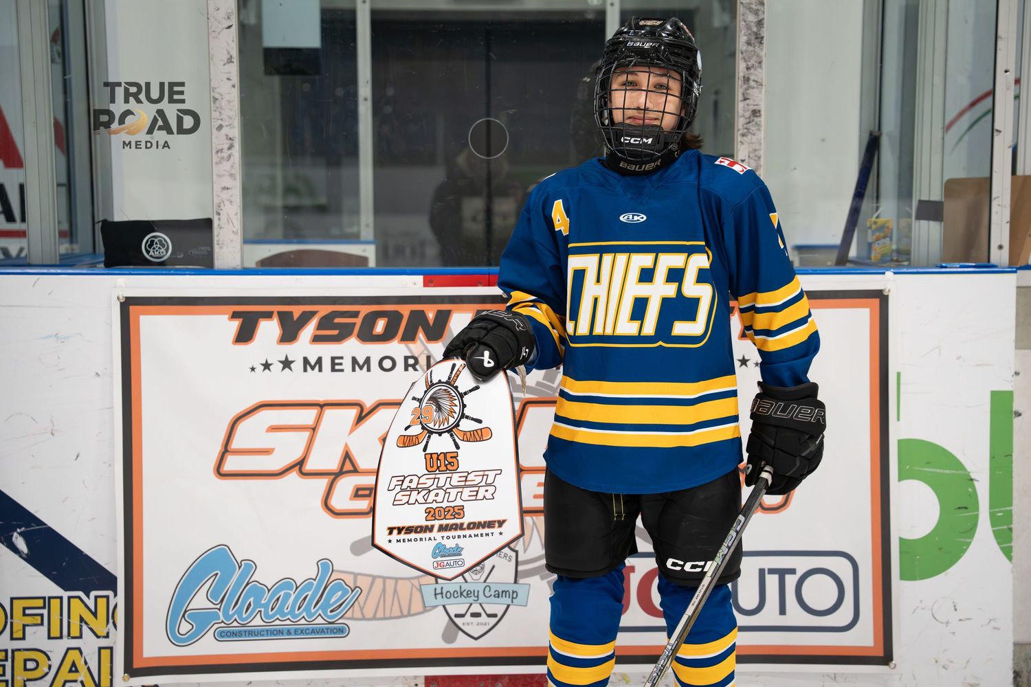 A youth hockey player in a blue and yellow Chiefs jersey stands on the ice rink in front of advertising boards.