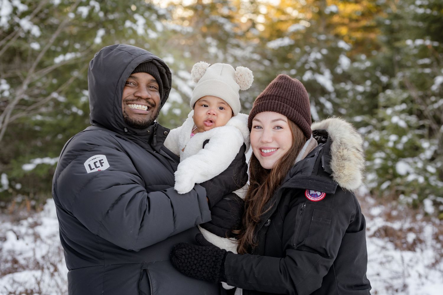 A family in winter coats smiles together in a snowy forest setting during golden hour.